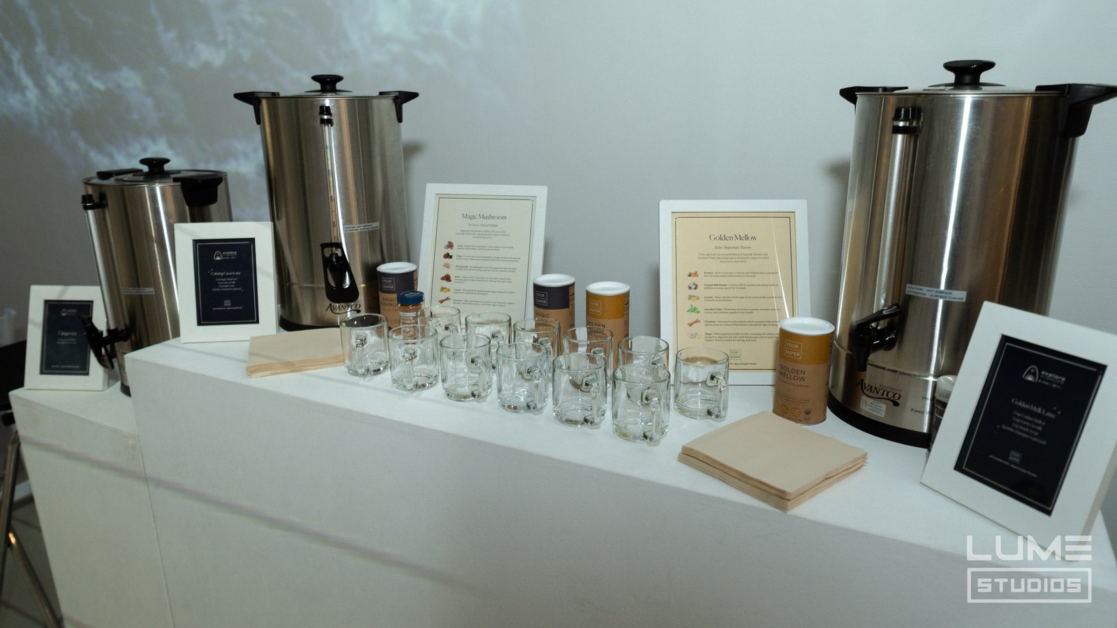 Steel hot water urns, glass mugs, tea tins and framed menu cards arranged on a white counter