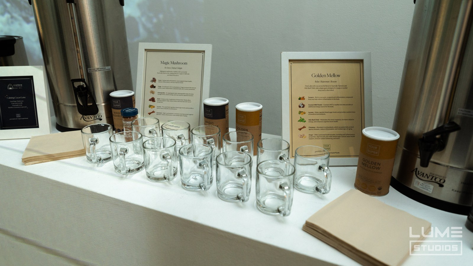 Rows of glass mugs and tea canisters between two steel urns with framed menu cards behind