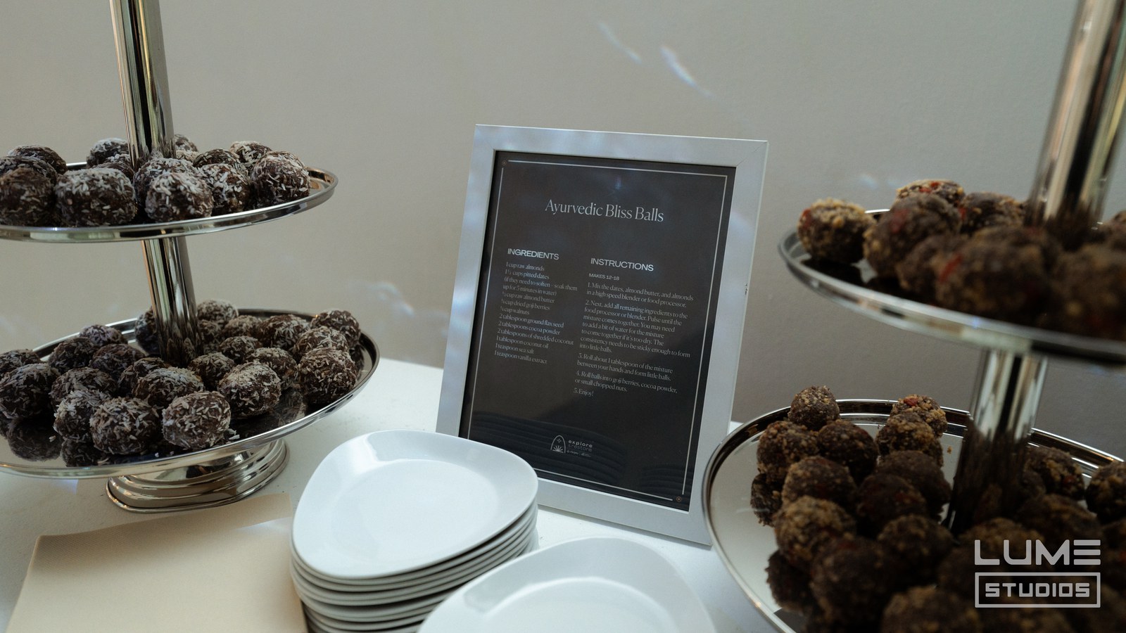 Tiered silver stands of coconut dusted bliss balls beside stacked white plates and a framed recipe card
