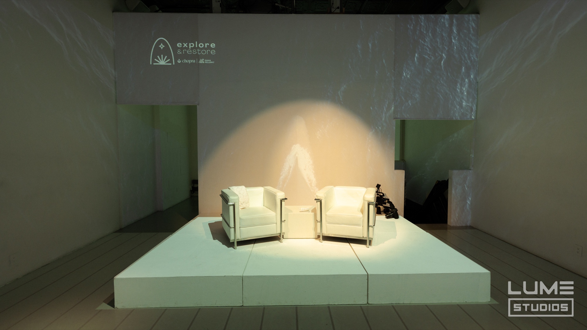 Two white armchairs on a low white stage lit by a warm spotlight, an Explore logo projected above
