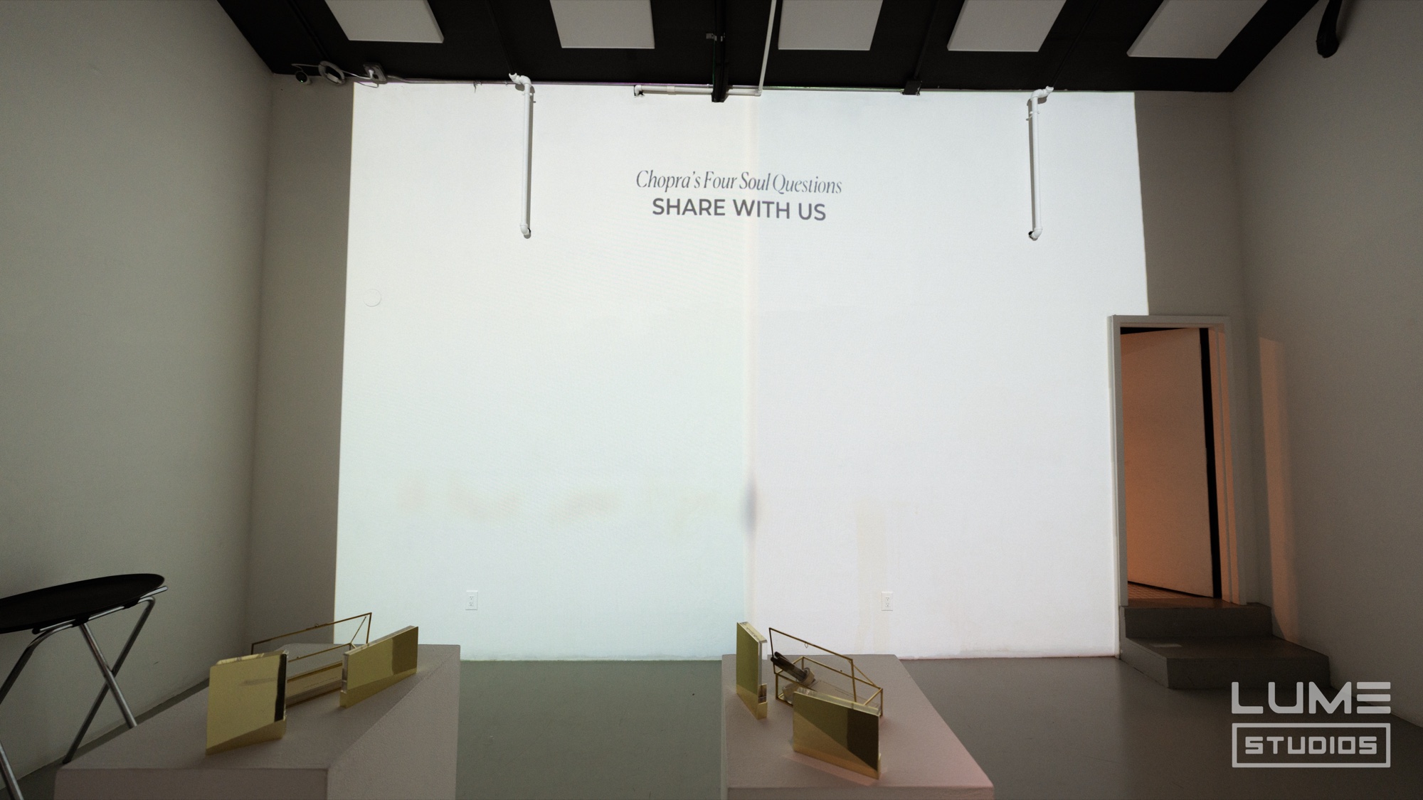 Bare white studio wall projected with the words Share With Us above two small brass frames on plinths