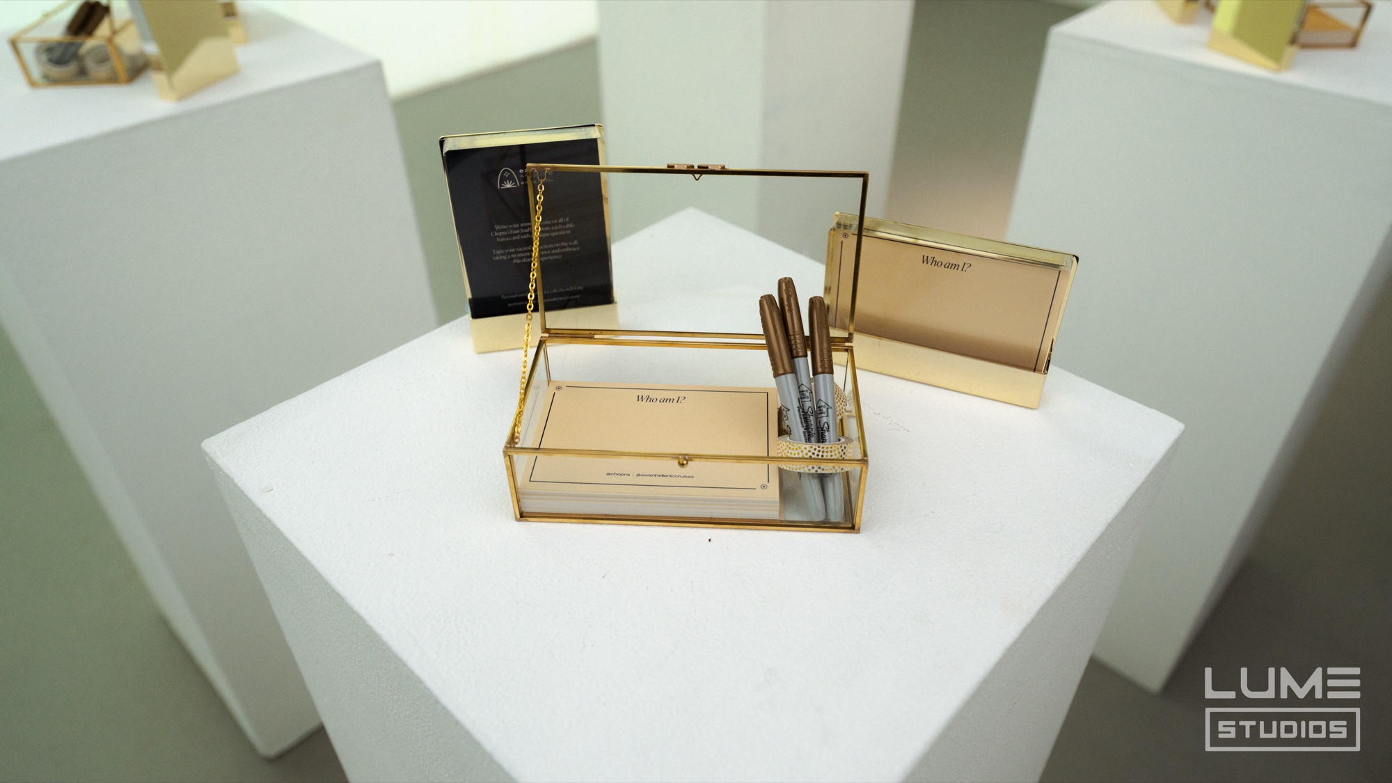 Gold framed glass box holding pens and cards on a white plinth, more gold boxes on plinths behind