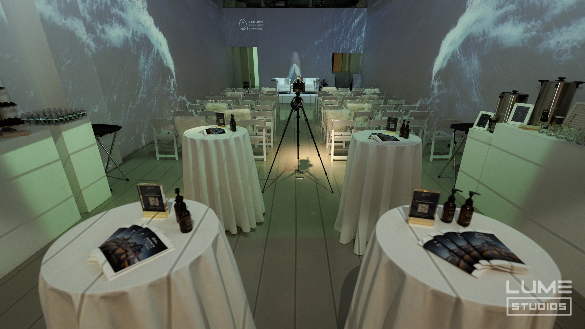 Round white tables with brochures and bottles face rows of white chairs under wave projections on the walls