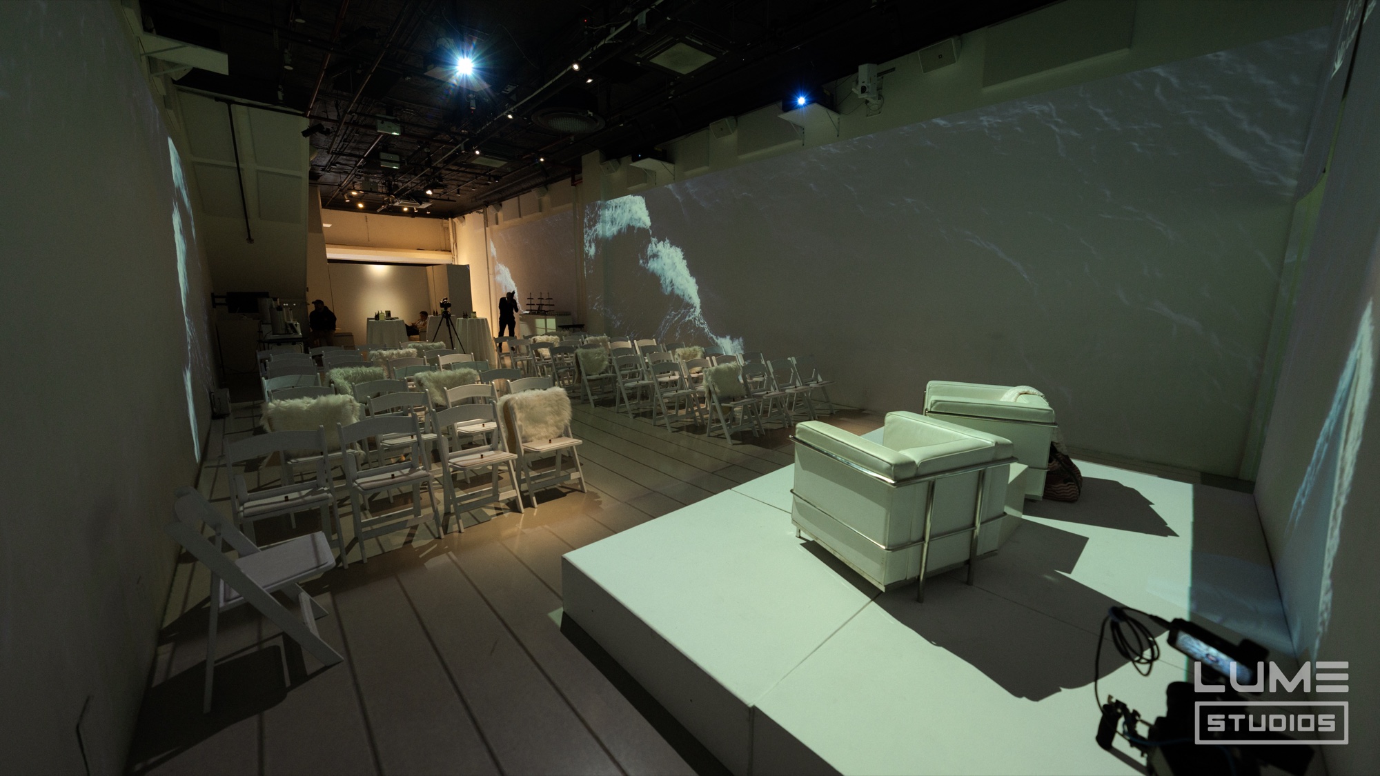 White armchair on a raised platform beside rows of white folding chairs under a rippling light projection