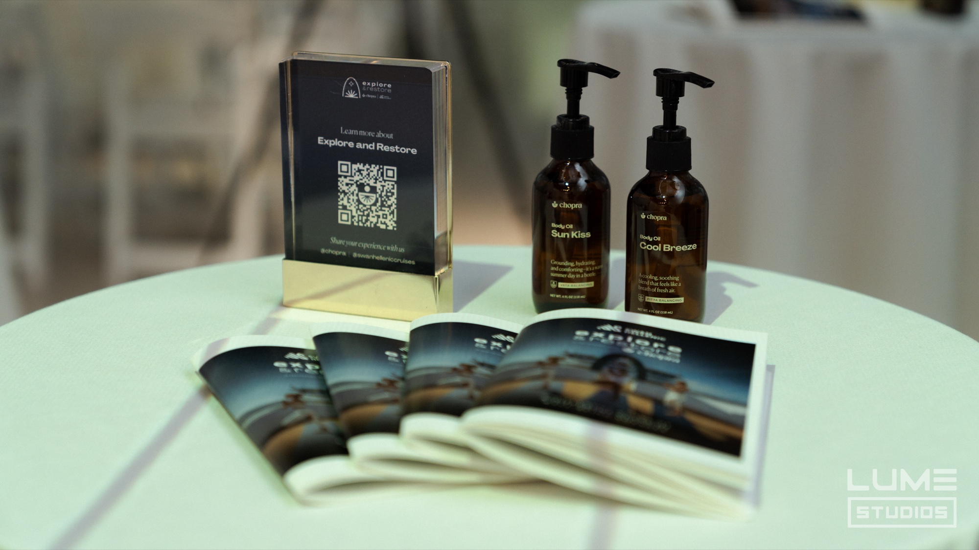 Two amber pump bottles, a Chopra QR code card and fanned brochures on a pale green table