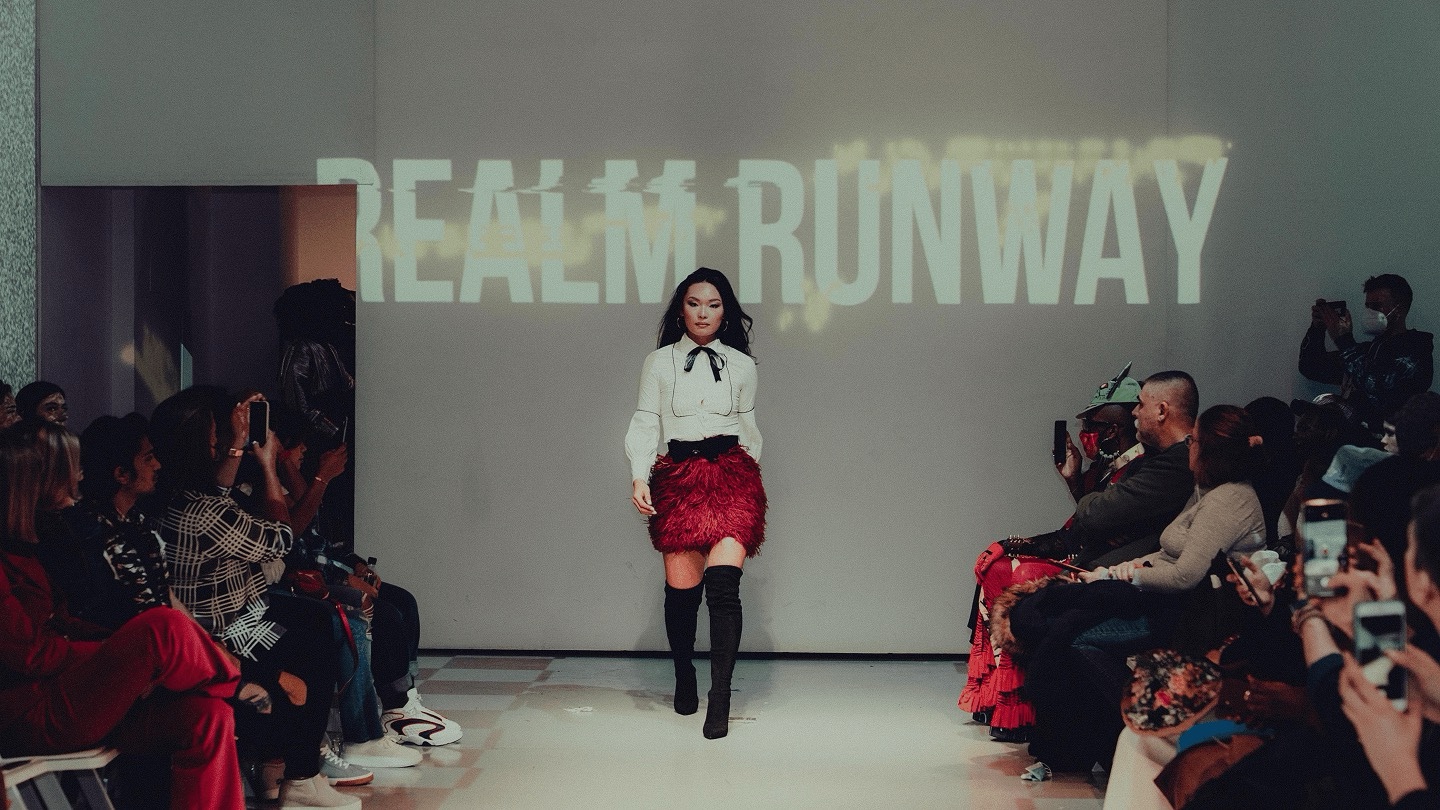 Model walks the DigitalAX show in a red feathered skirt under a Realm Runway projection