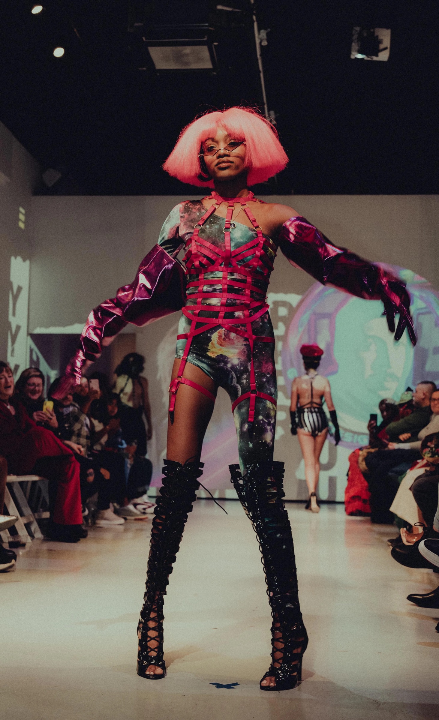Model in a pink bob wig, red harness top and lace up boots poses at the end of the runway