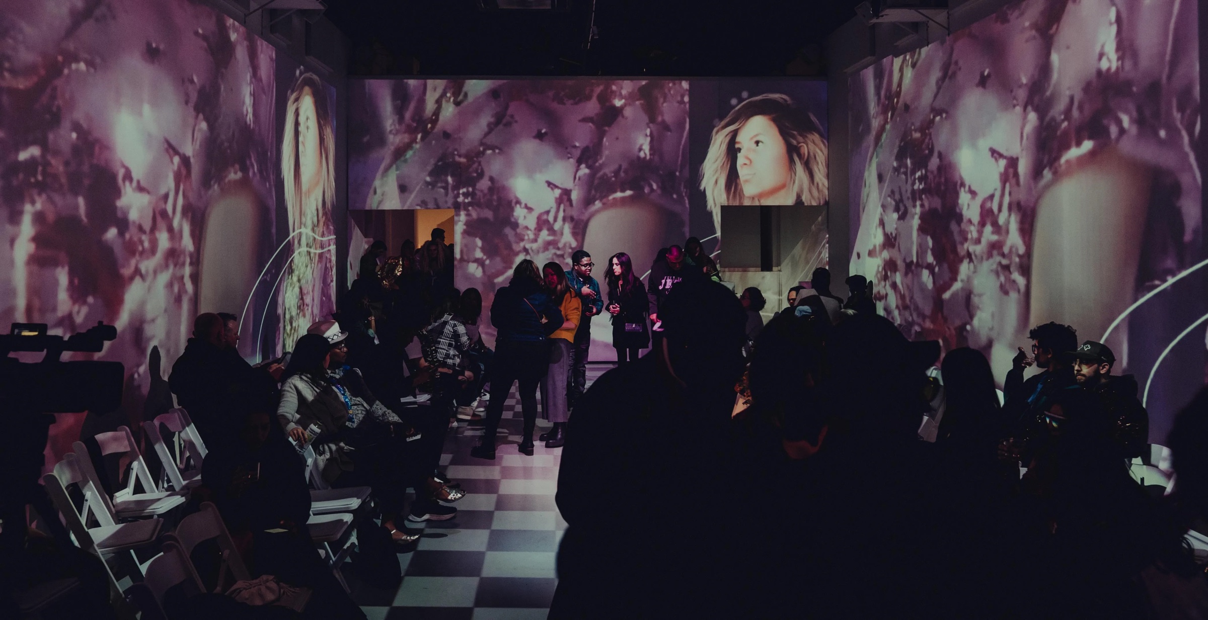 Wide view of the DigitalAX runway room with projected artwork on the walls and a seated audience on both sides