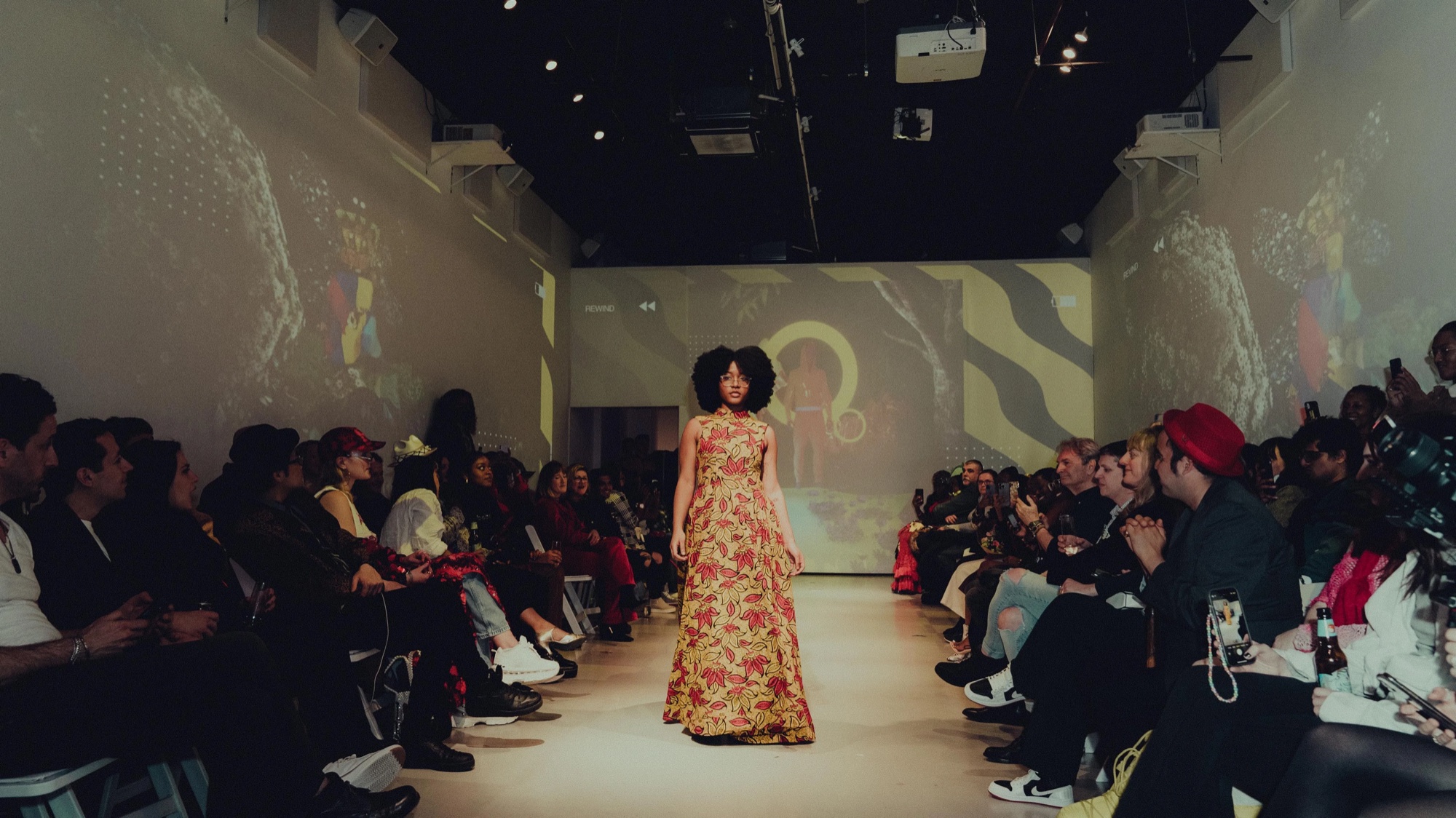 Model in a gold floral gown walks the DigitalAX runway past a full audience at Lume Studios