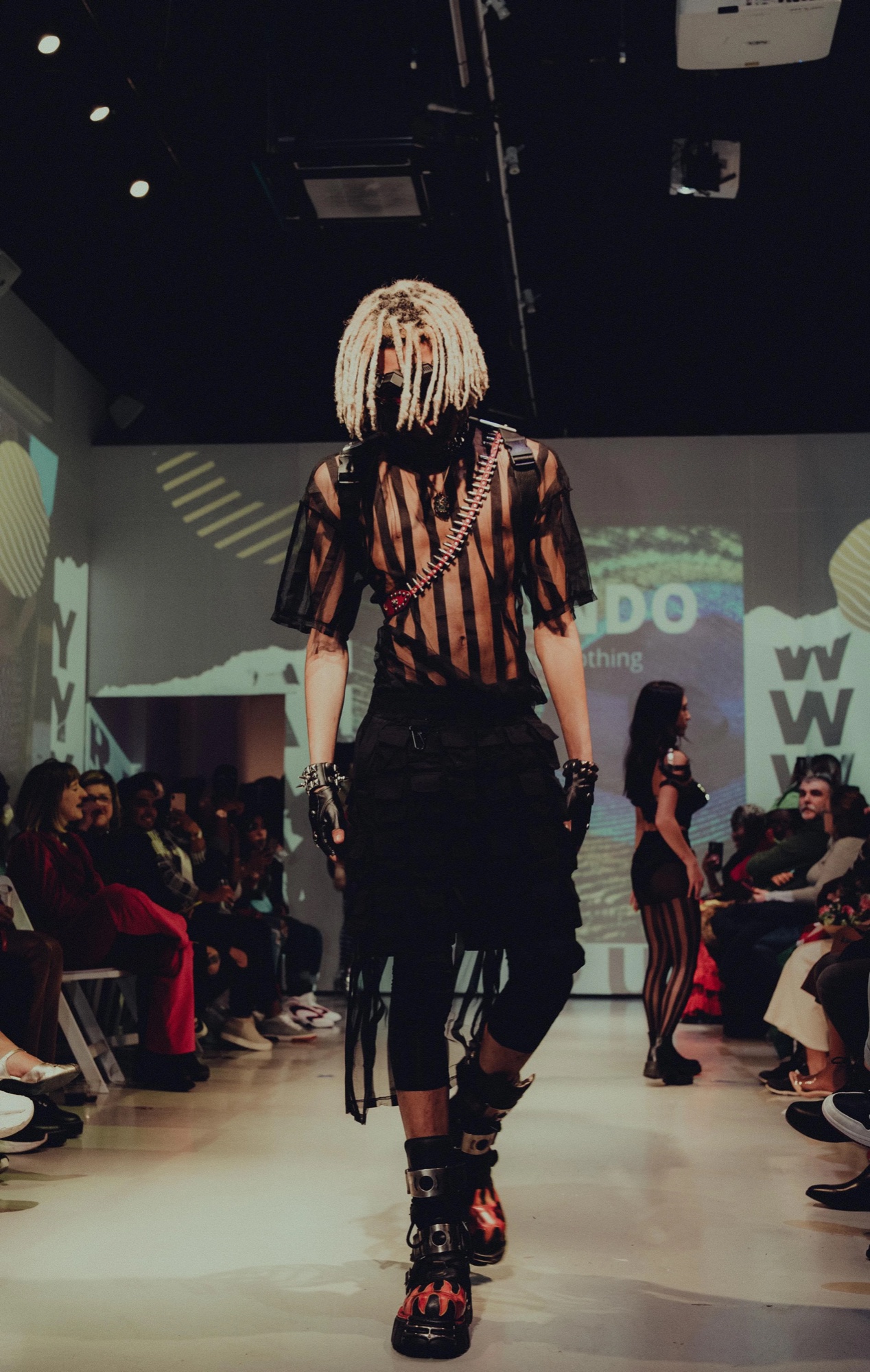 Model in a braided silver mask and sheer black look walks the runway under projected graphics