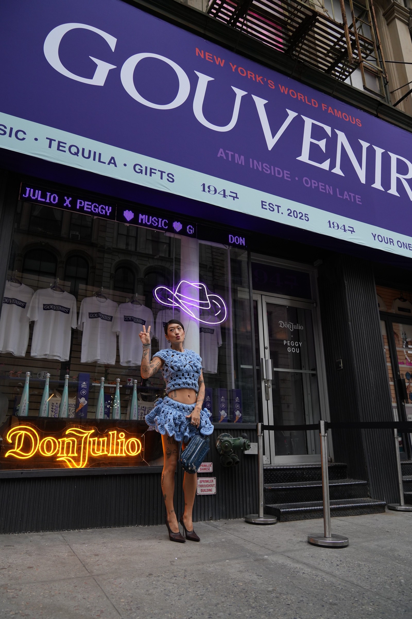 A woman in a blue crochet dress poses outside a storefront with a Don Julio neon sign and Gouvenirs banner