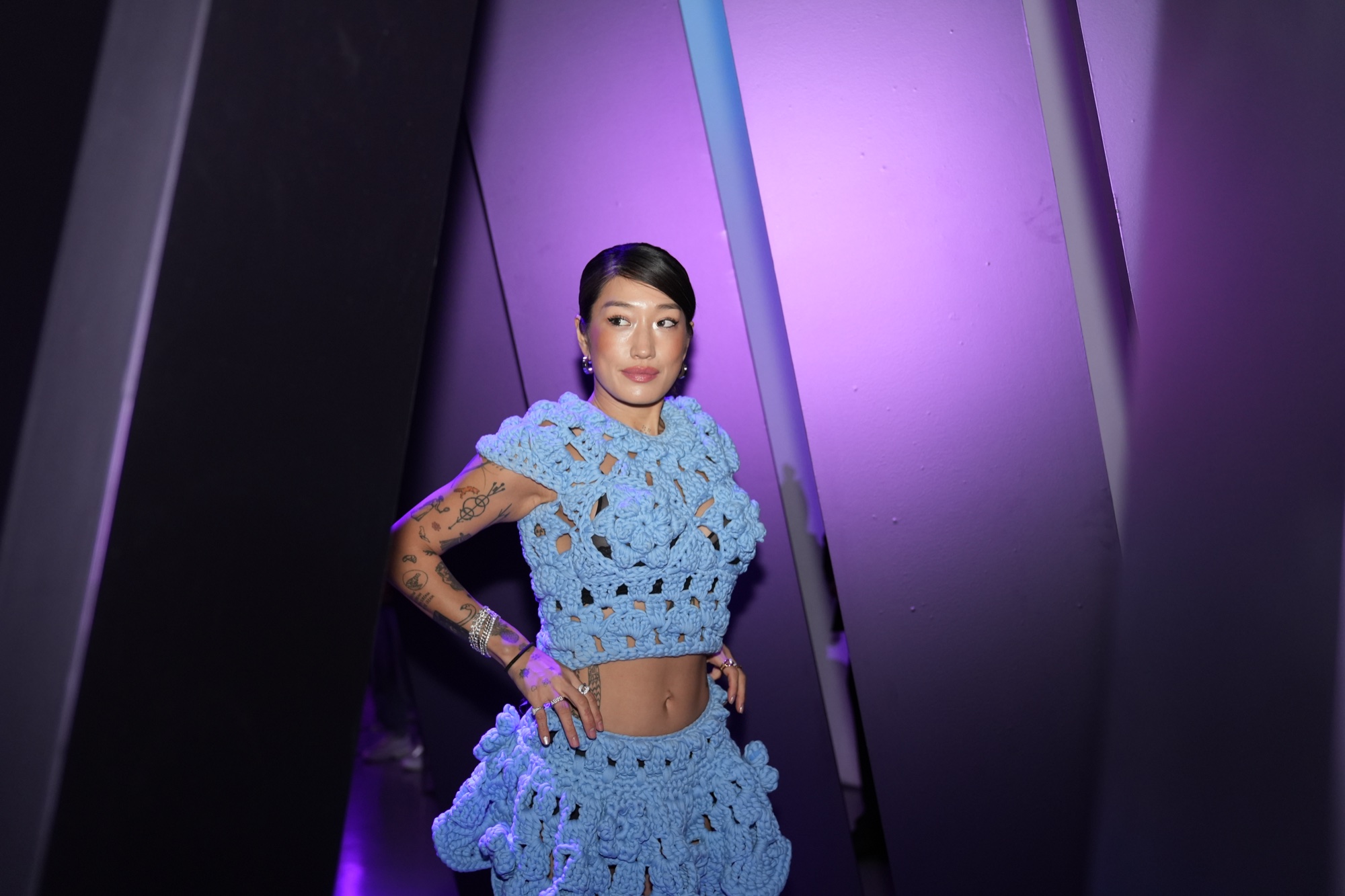 Woman in a light blue crochet top and skirt posing hands on hips between purple lit angled panels