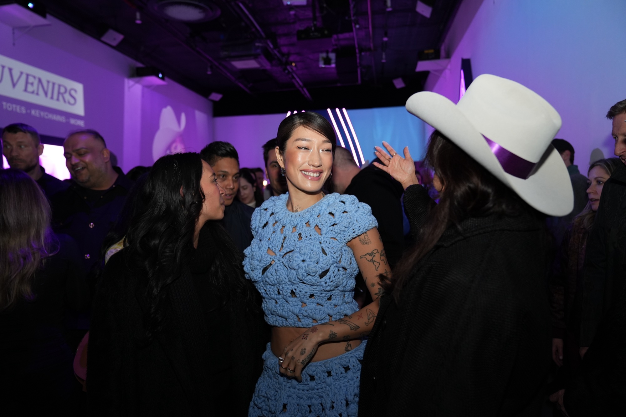 A woman in a blue crochet dress laughs in a crowd beside a guest in a white cowboy hat