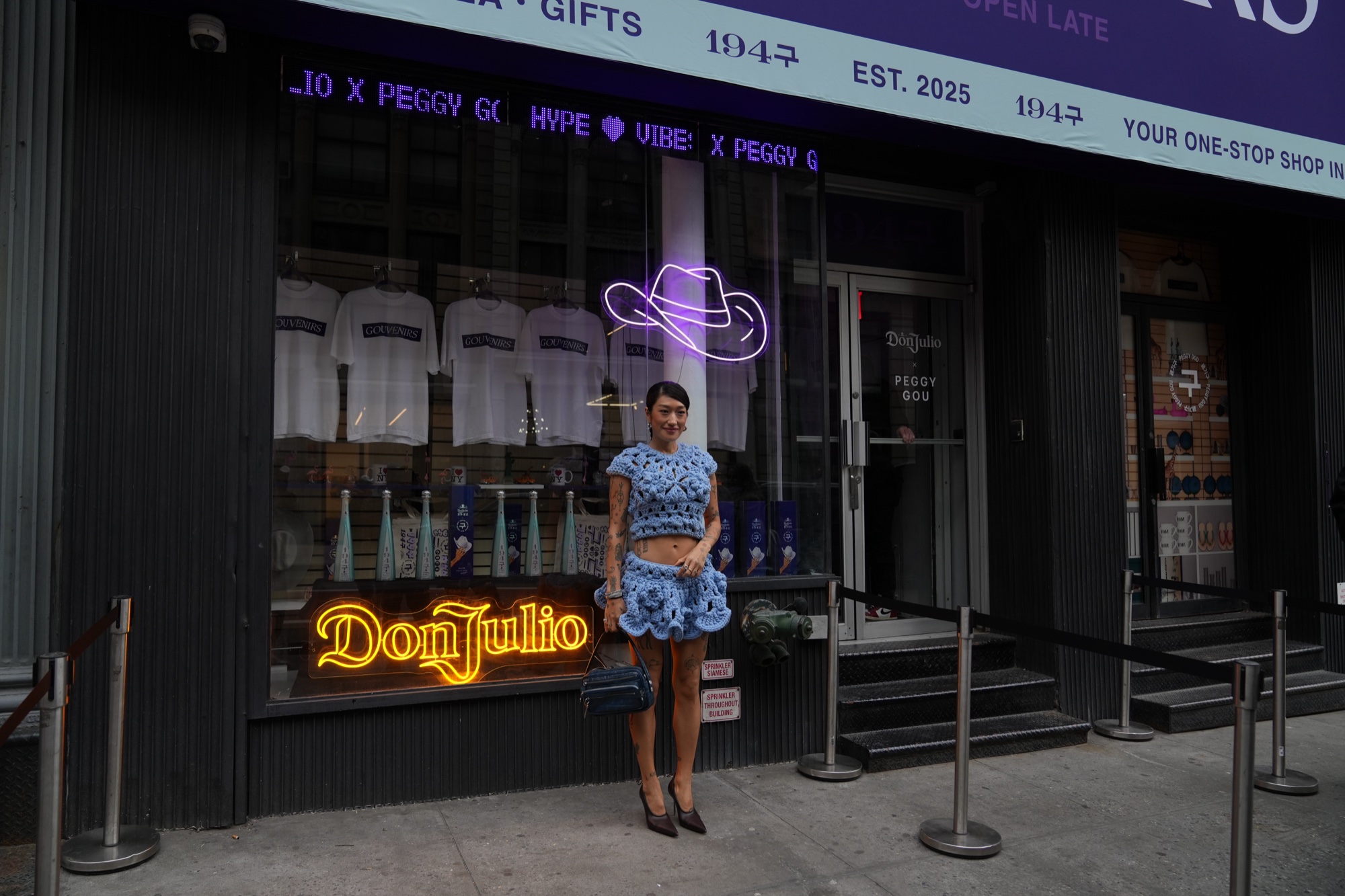 A woman in a blue crochet dress stands before a bodega storefront with an orange Don Julio neon sign