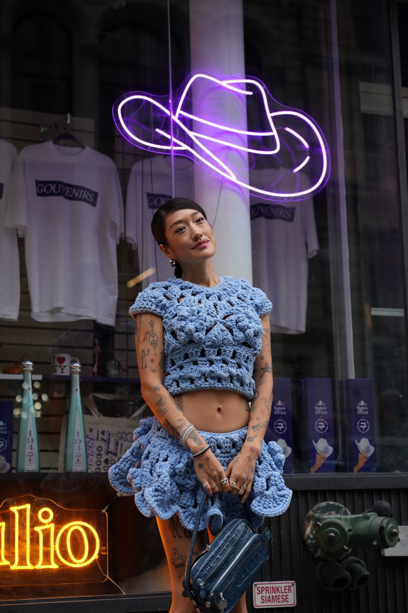 Sideways frame of a person in a blue crochet top and skirt beside a purple neon figure in a window