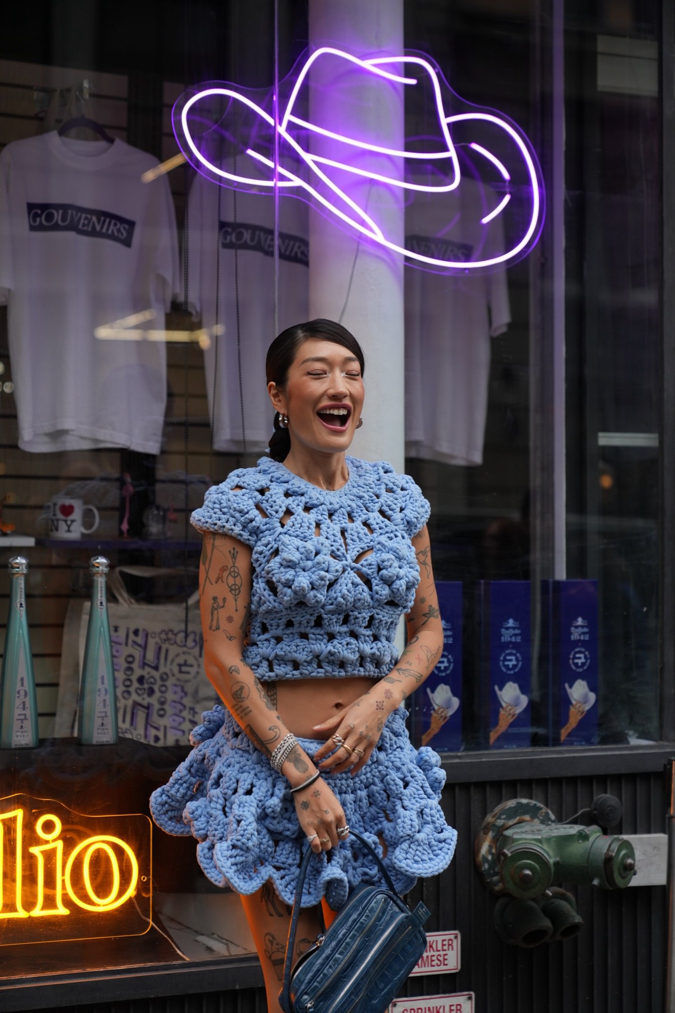 Rotated view of a laughing woman in a blue crochet outfit beside a purple neon outline in a shop window