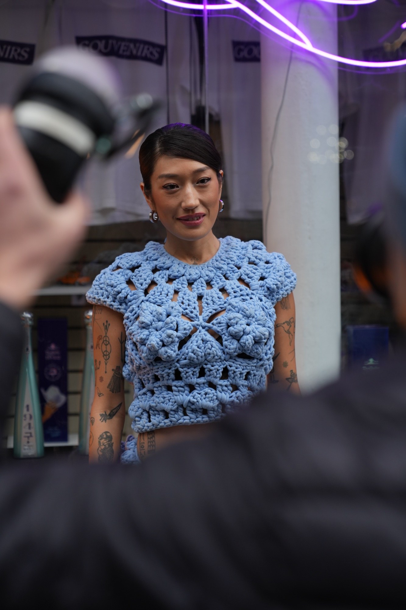A tattooed woman in a blue crochet top is photographed beside a purple neon shape inside a merchandise shop