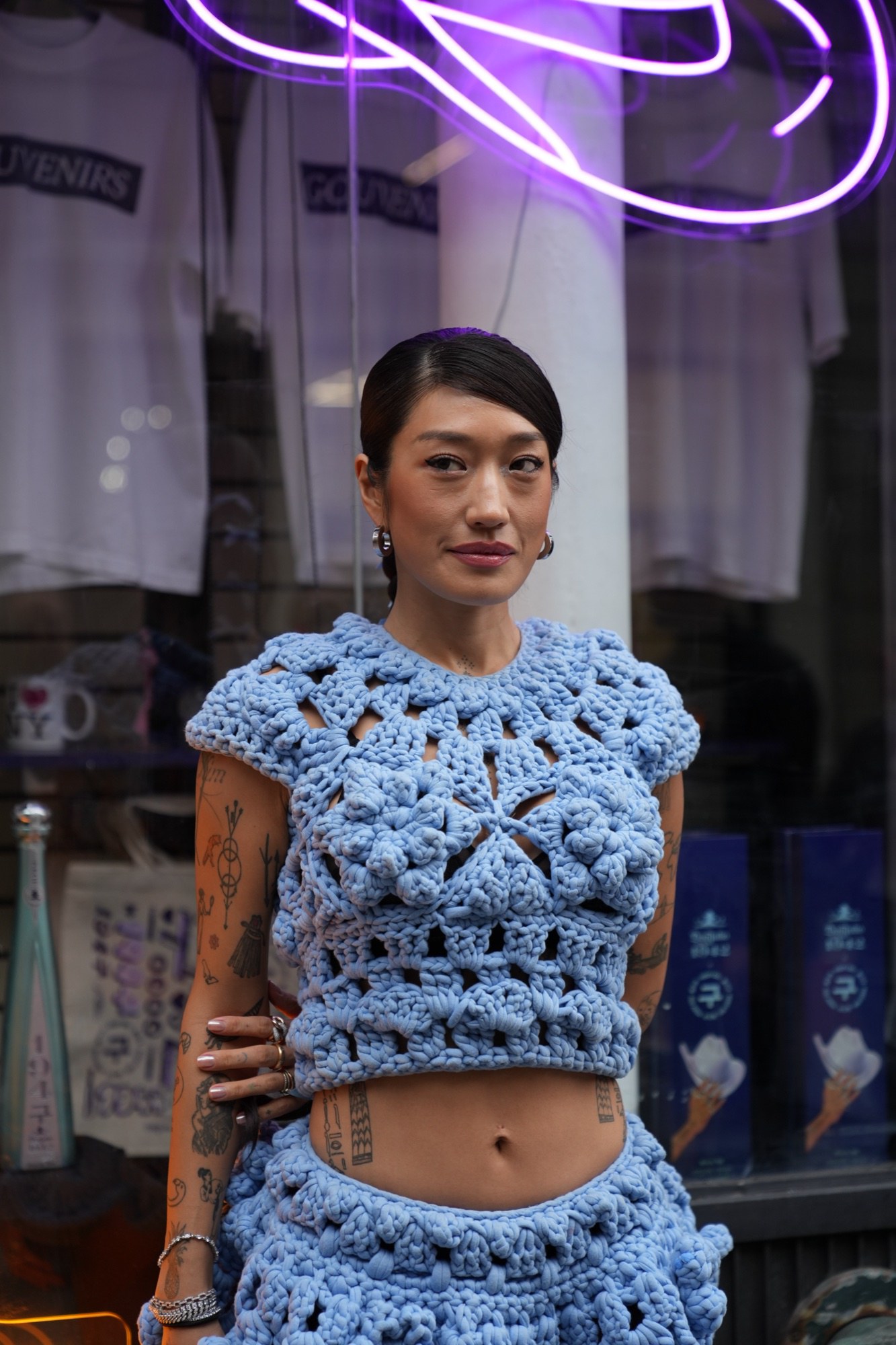 A tattooed woman in a blue crochet two piece stands in a store aisle beside a purple neon sign