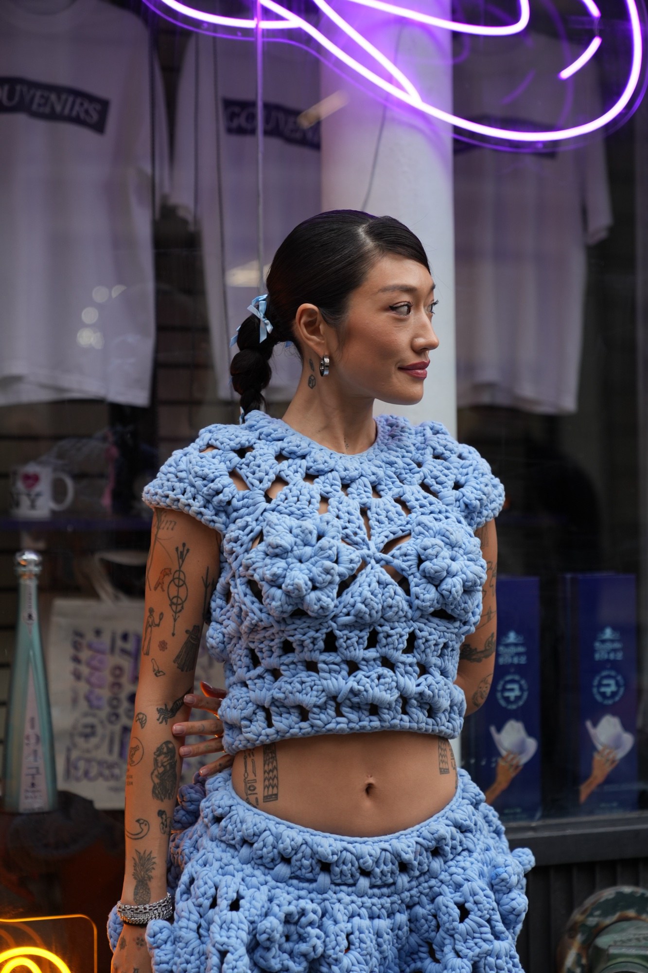Rotated close view of a tattooed woman in a blue crochet two piece looking down past a purple neon