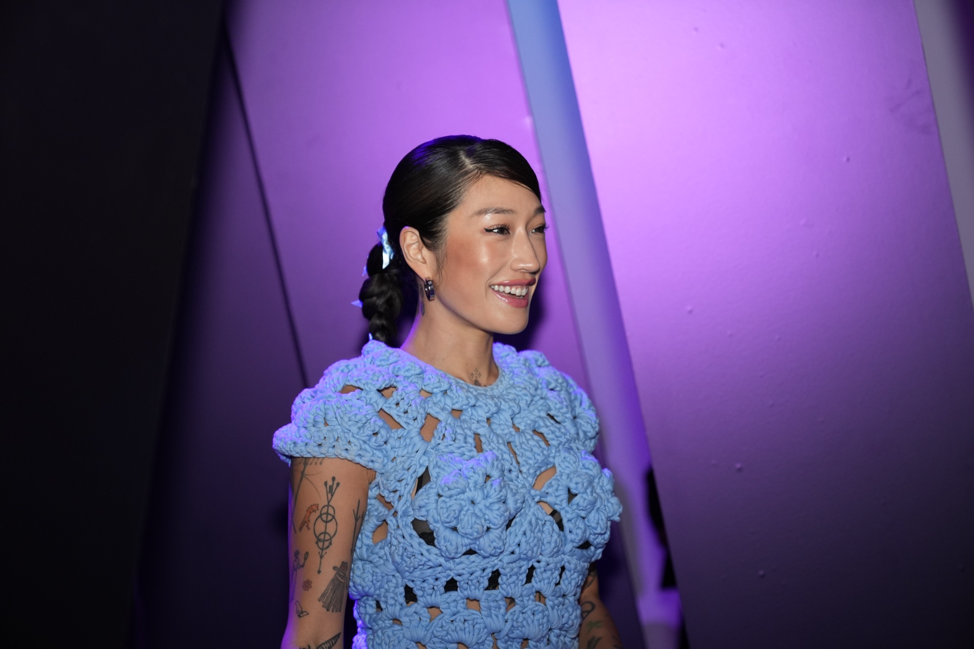 A smiling woman in a blue crochet dress with tattooed arms stands in purple and lilac light