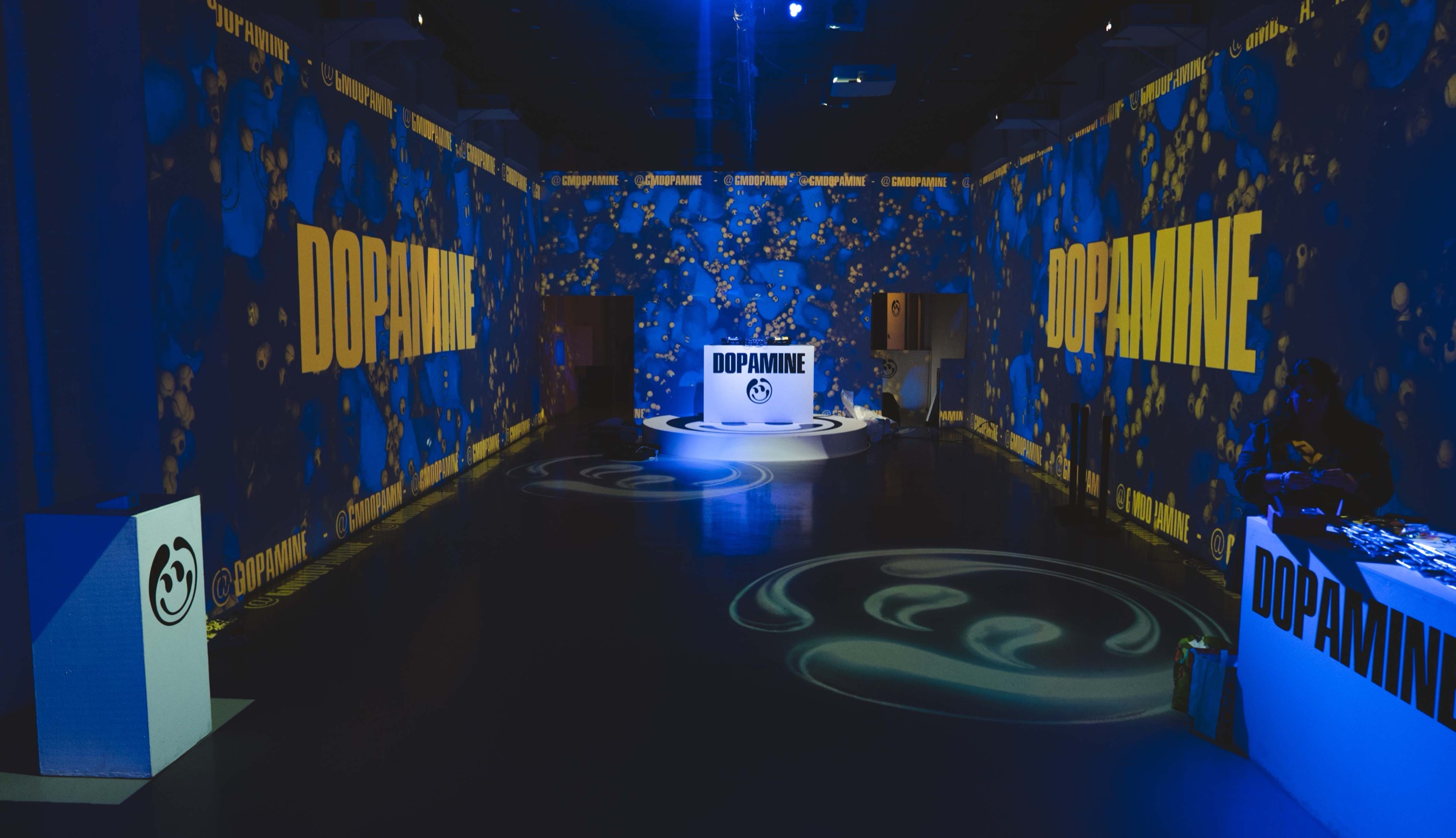 Dark event space washed in blue with yellow Dopamine projections and a glowing DJ booth