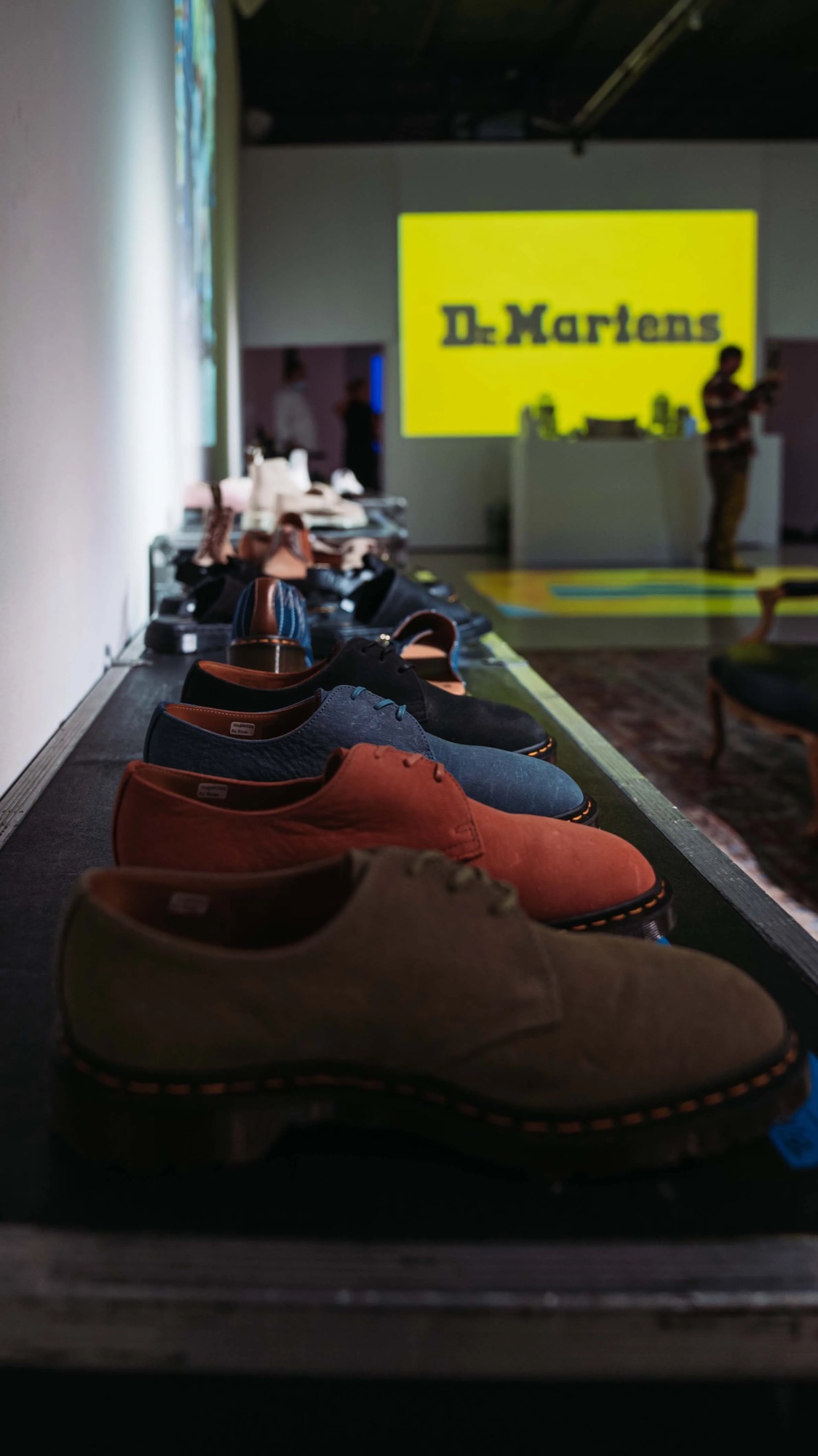 Row of suede Dr. Martens shoes on a road case with the yellow logo projected behind