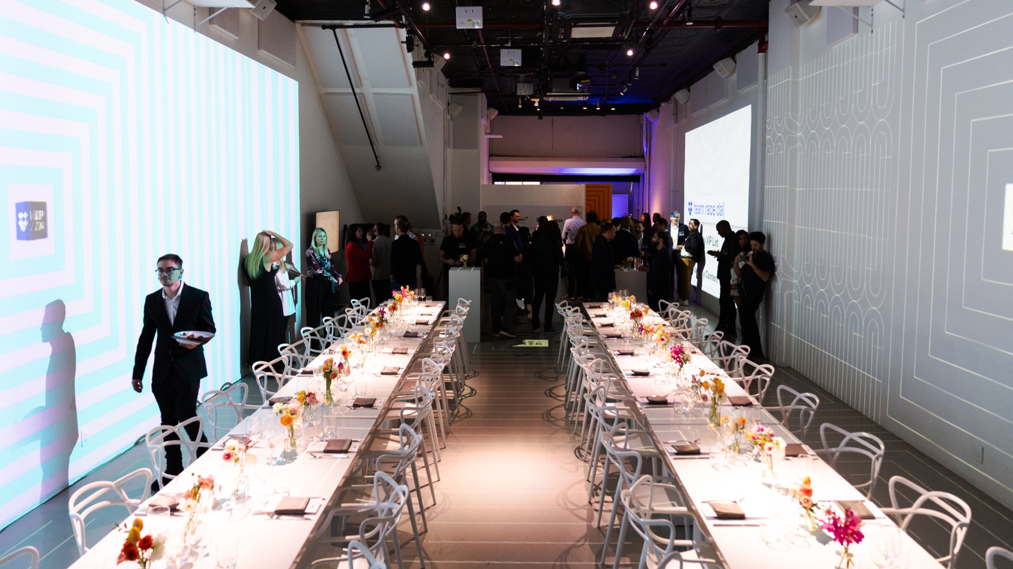 Two long white dinner tables lined with wire chairs and flowers in a hall with striped projection walls
