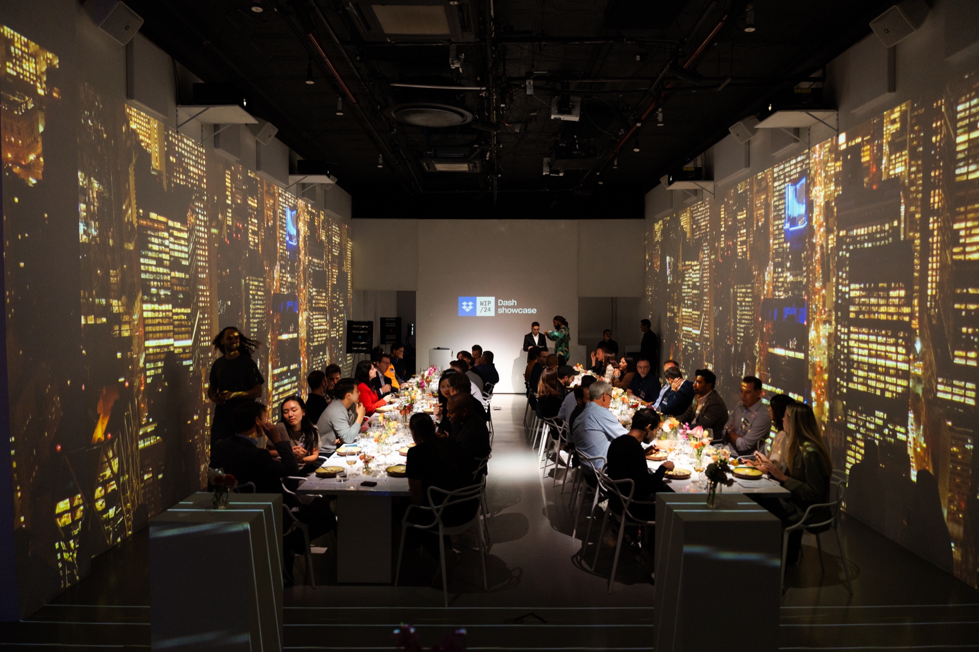 Guests dine at long tables in a dark room while glowing city skylines are projected across both walls