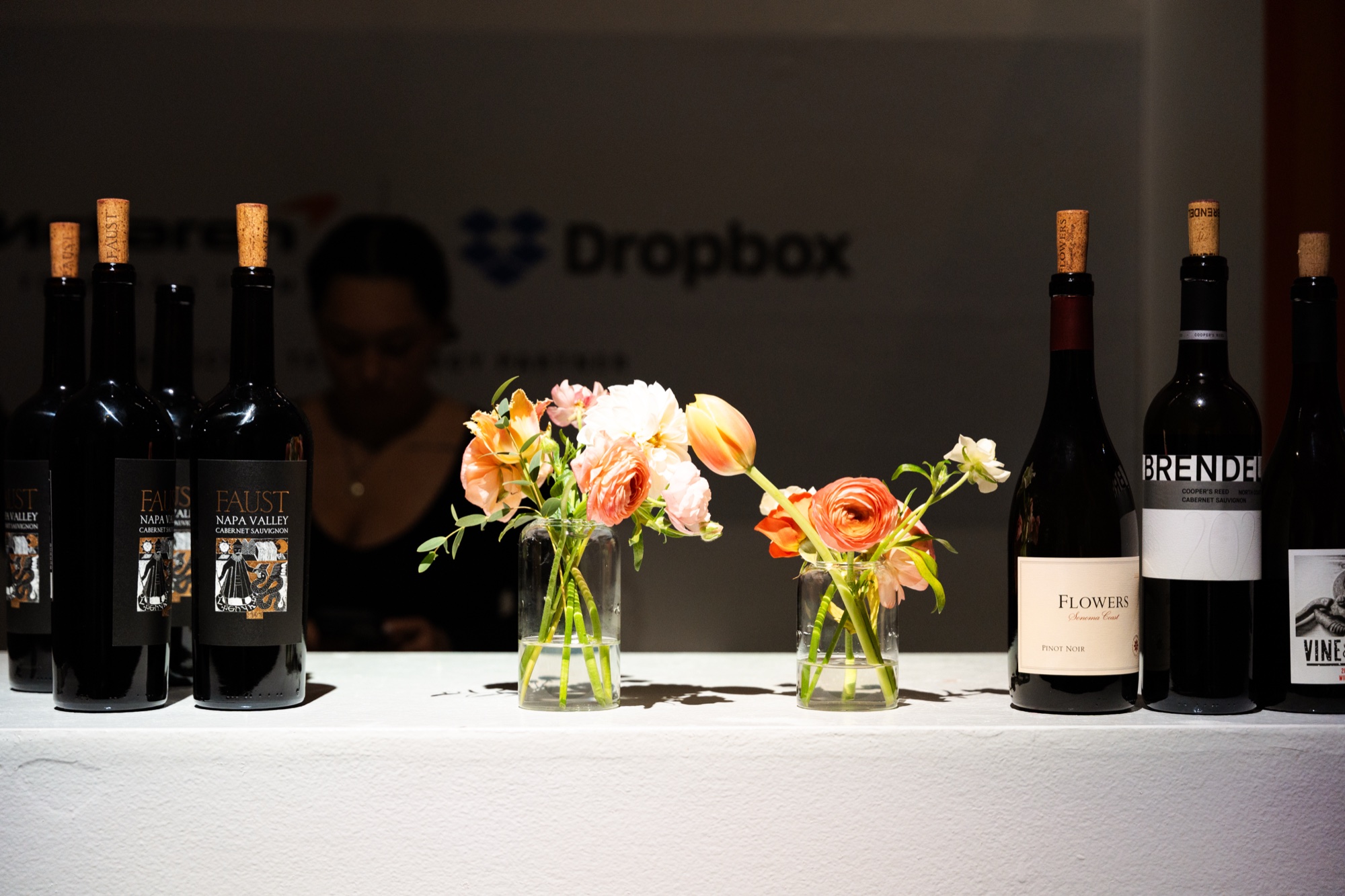 Red wine bottles and small vases of orange and pink flowers on a white bar beneath a Dropbox logo