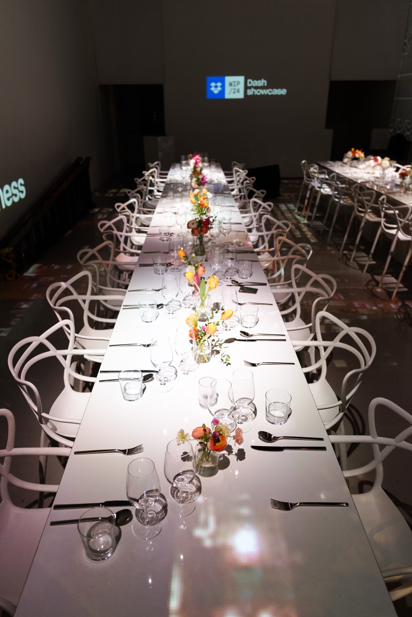 A long white table set with glasses, cutlery and small orange and pink bouquets below a Dash showcase projection