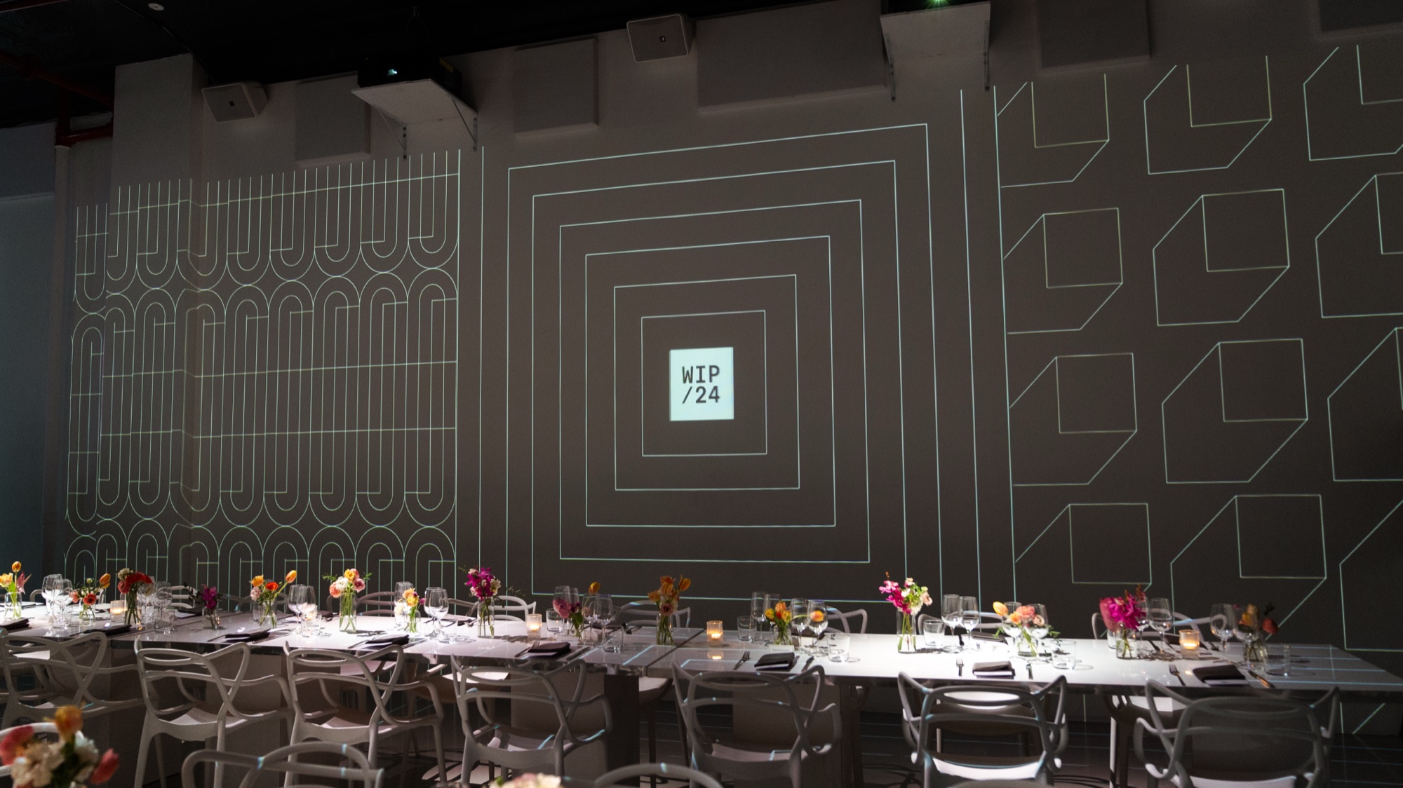 White geometric line patterns projected on dark walls above dinner tables set with bright flowers, WIP/24 at centre