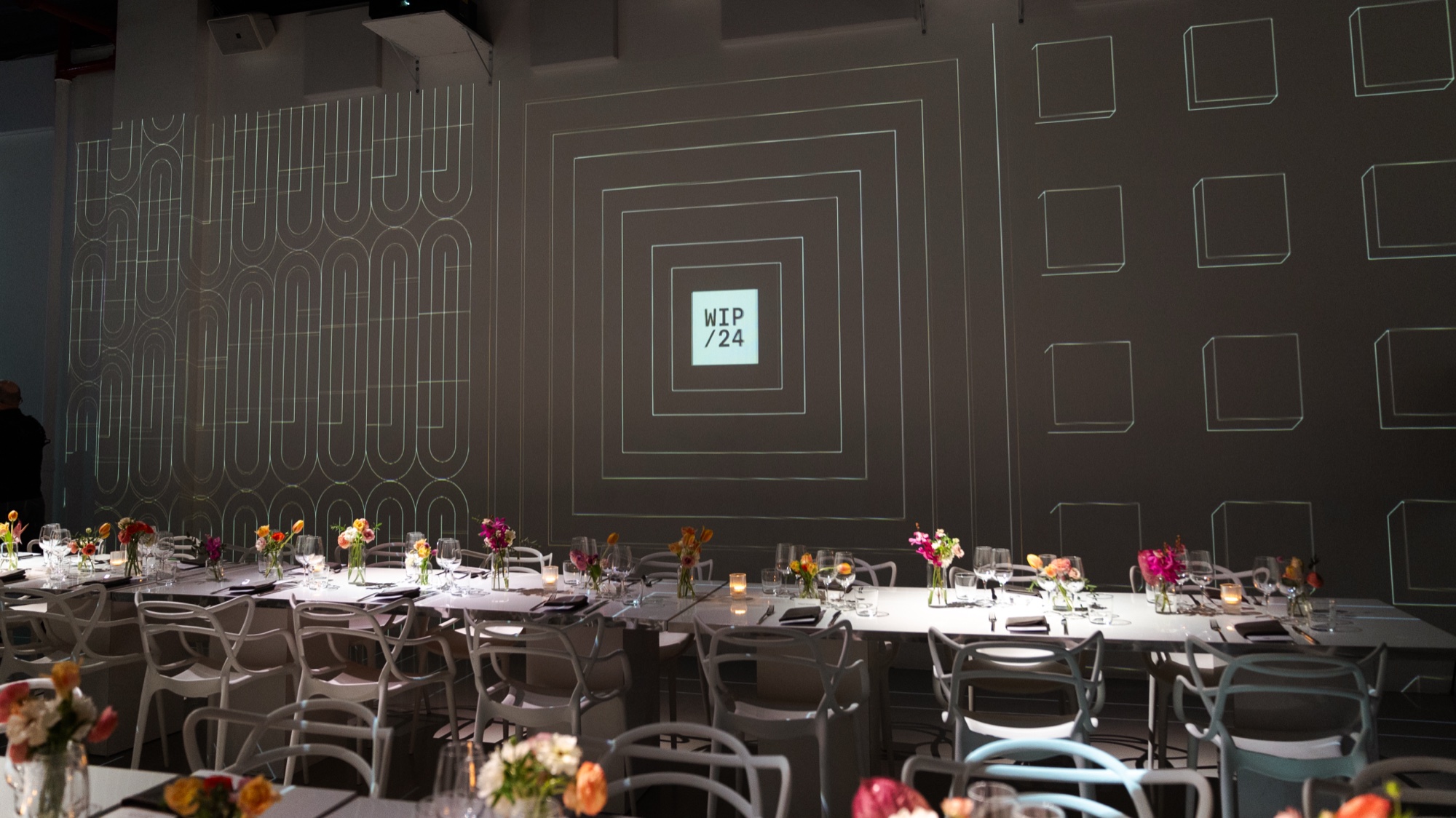 Round tables with flowers and glassware before a dark wall projected with white line patterns and a WIP 24 square