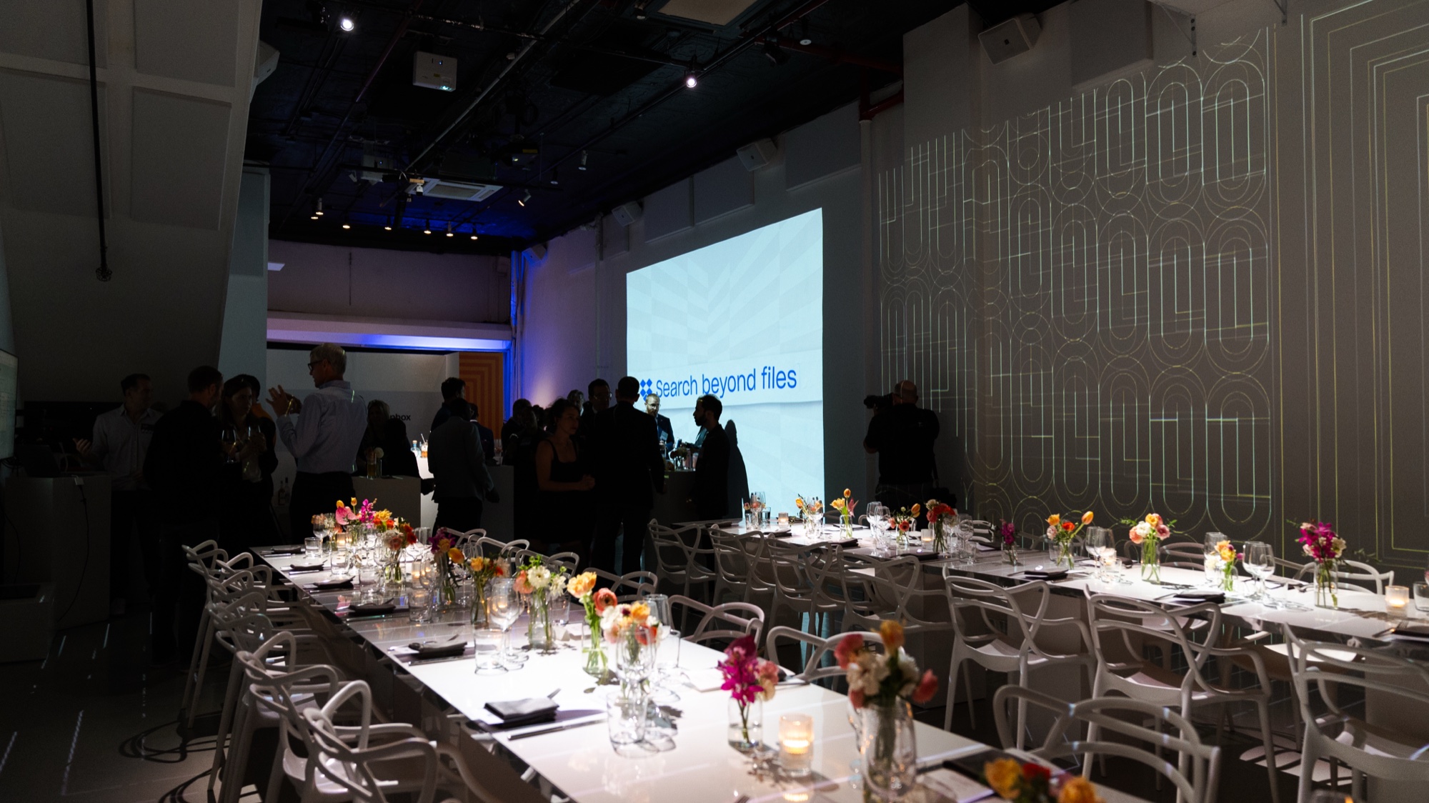 Guests mingling beside long white tables of flowers under a projected slide reading search beyond files