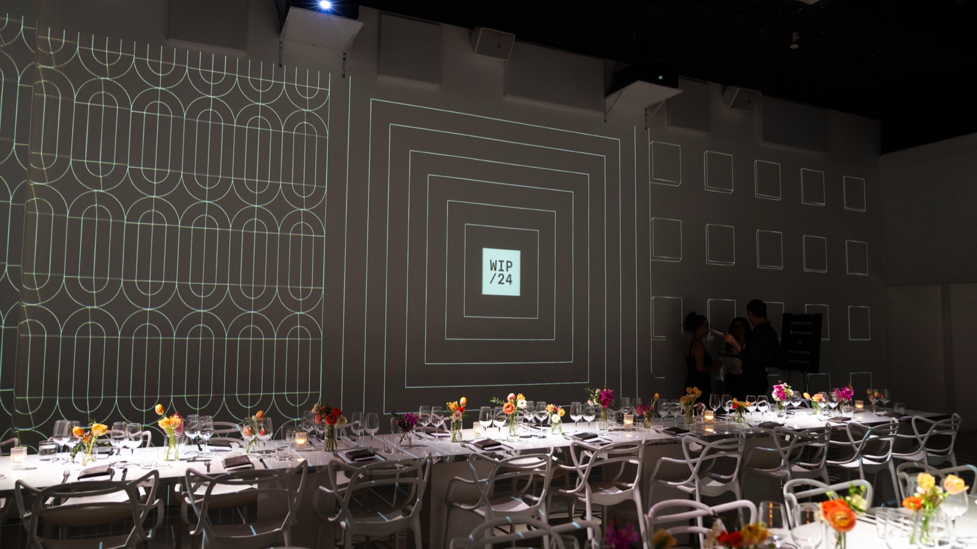 White line patterns and a WIP 24 badge are projected on a grey wall above tables set with bright flowers