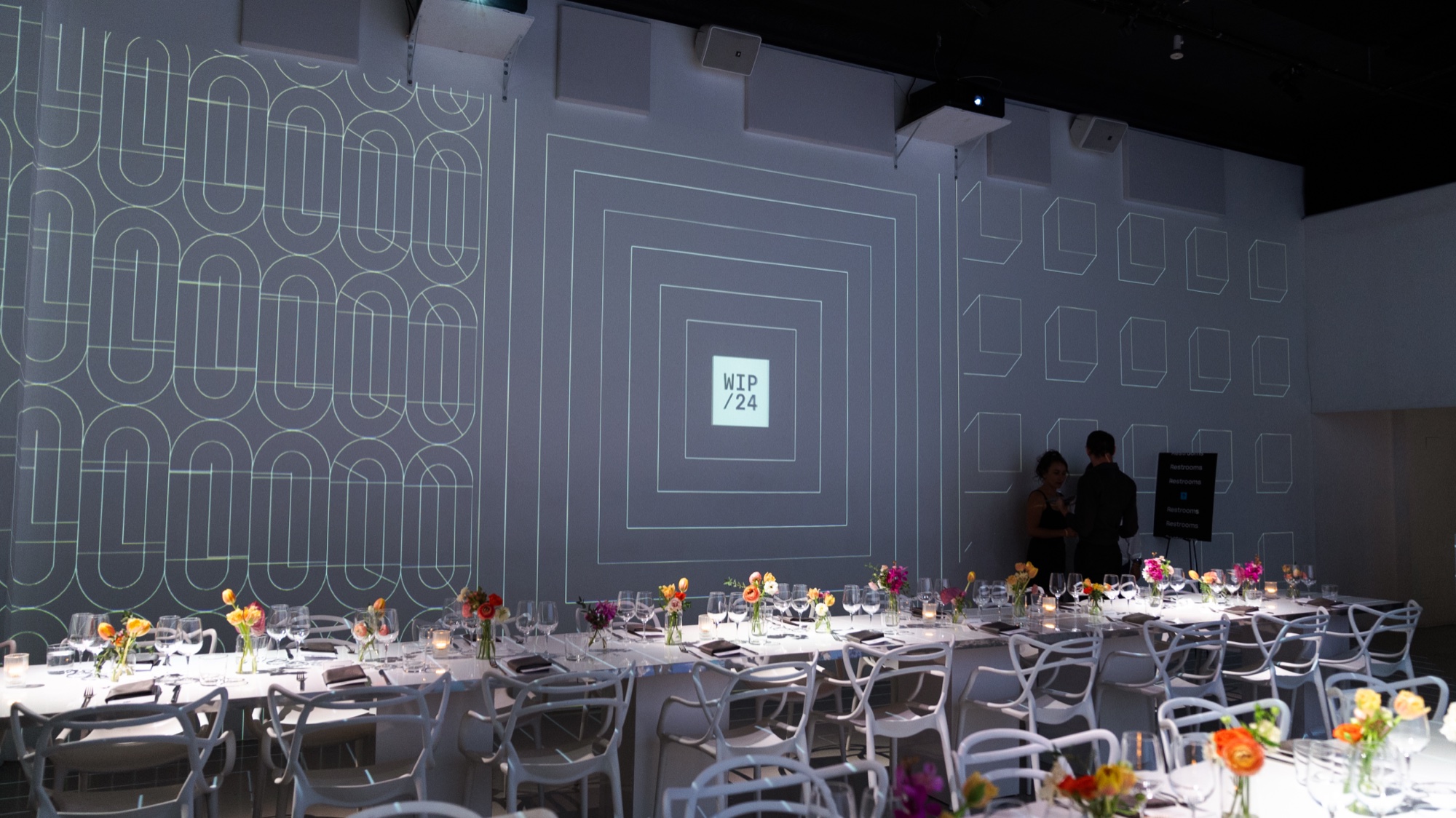Long tables with white chairs and colourful flowers under a wall of projected white geometric patterns reading WIP/24