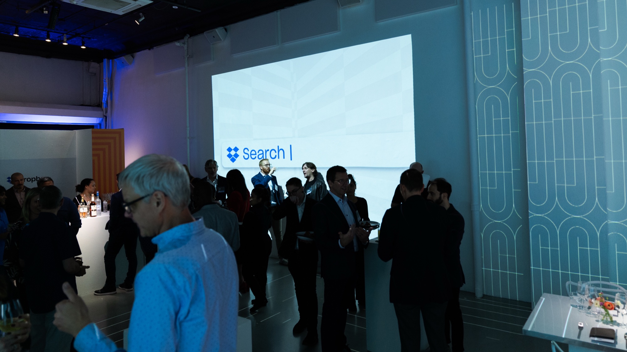 Guests mingle in a blue lit room beneath a large screen showing a Dropbox search bar