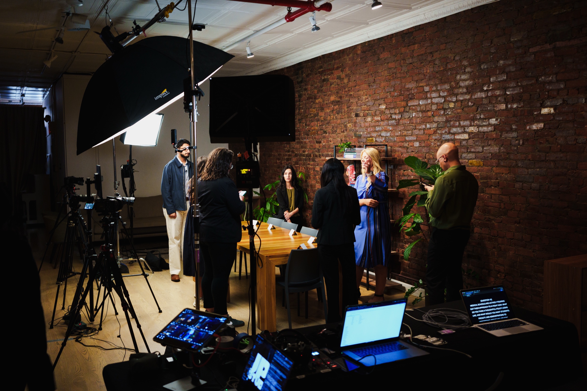 Crew and guests stand talking beside the wooden table as monitors and a softbox glow in the dark studio