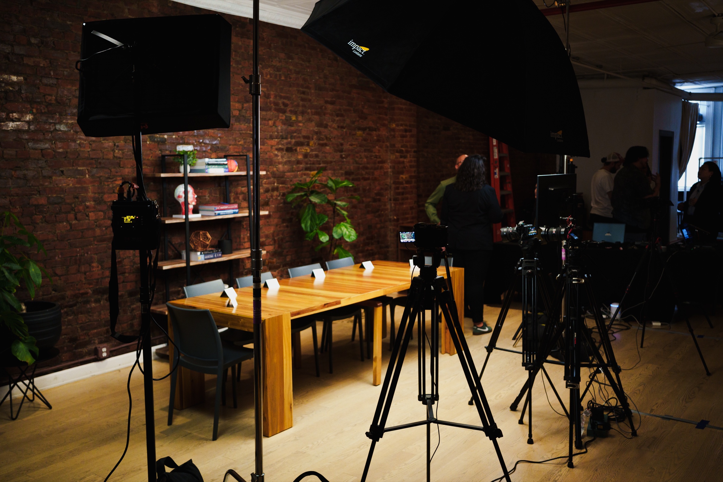 Empty set before taping, long wooden table lit warm against brick, cameras and umbrella light standing ready