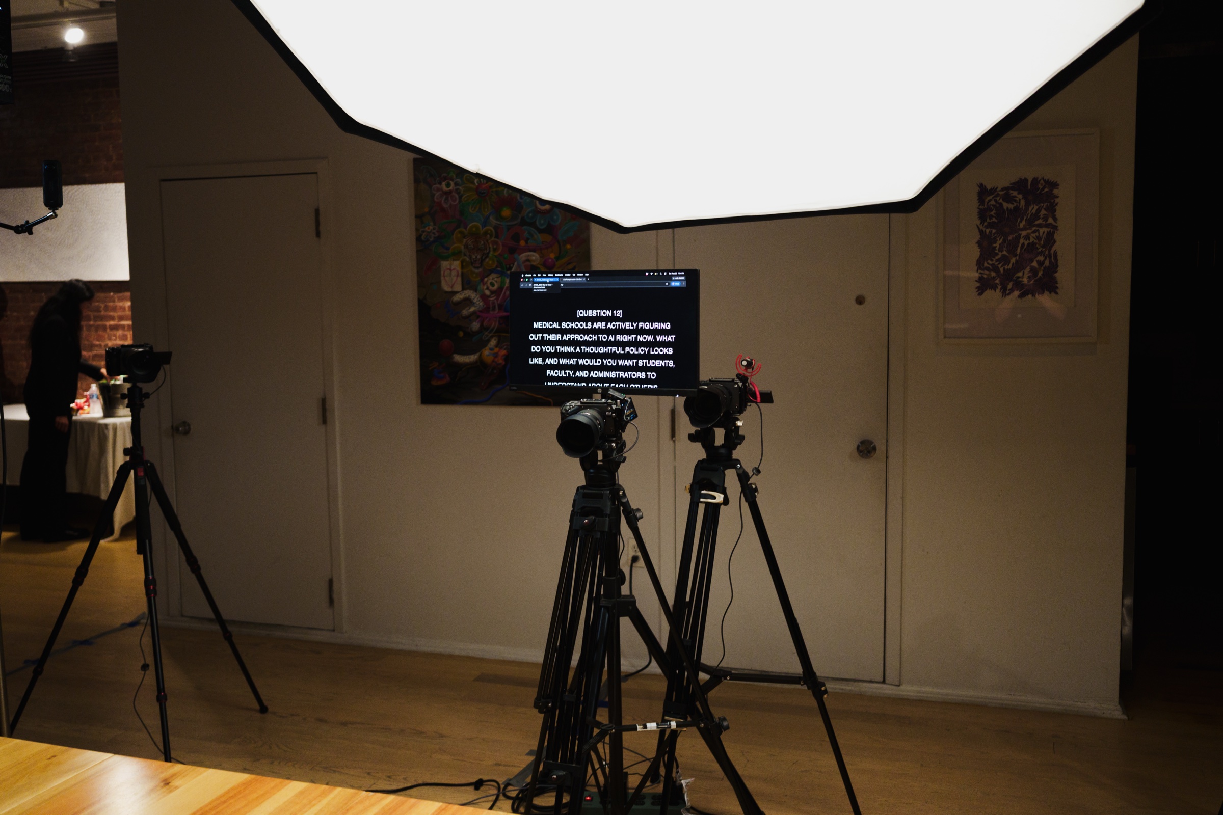 A wall monitor displays a written question behind two cameras under a large overhead softbox