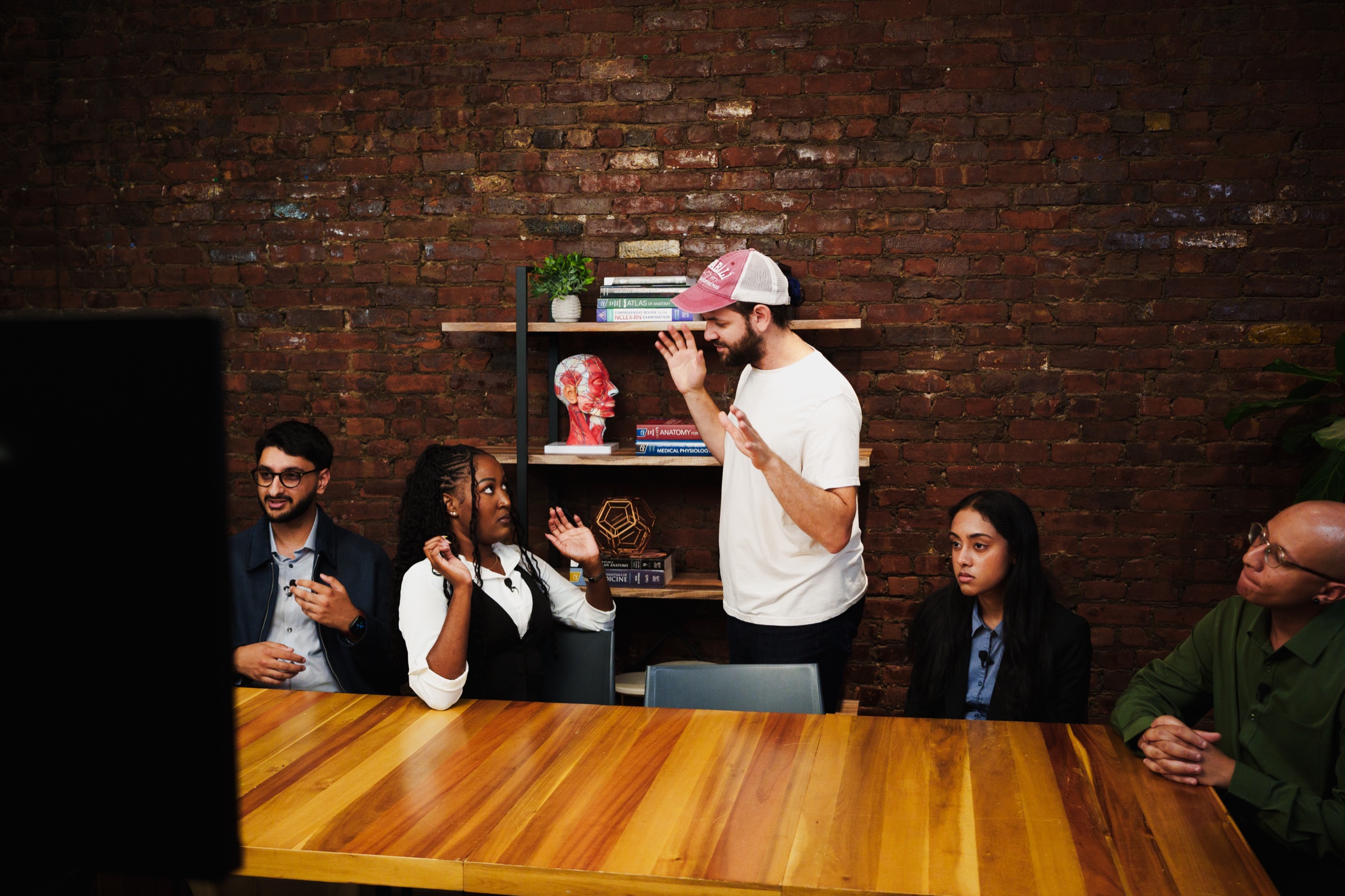 Doctor Mike stands in his pink cap gesturing as four seated guests listen along the wooden table