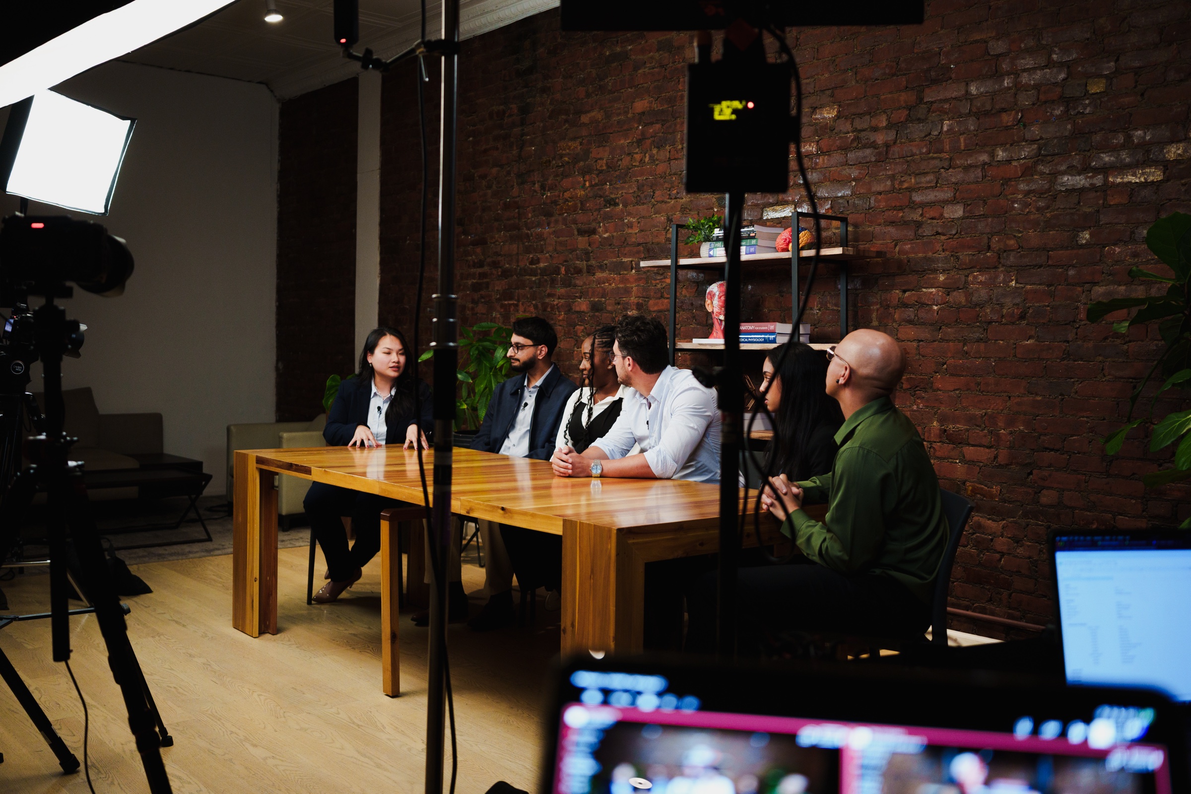 Four guests sit at the table talking with a man in green while a camera records in front