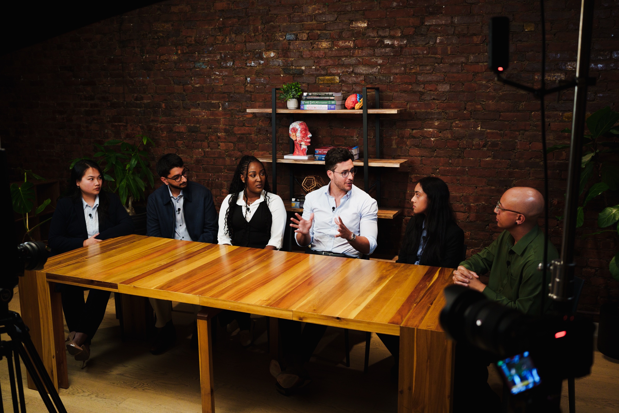 Six people sit along the long wooden table as the man in the center speaks