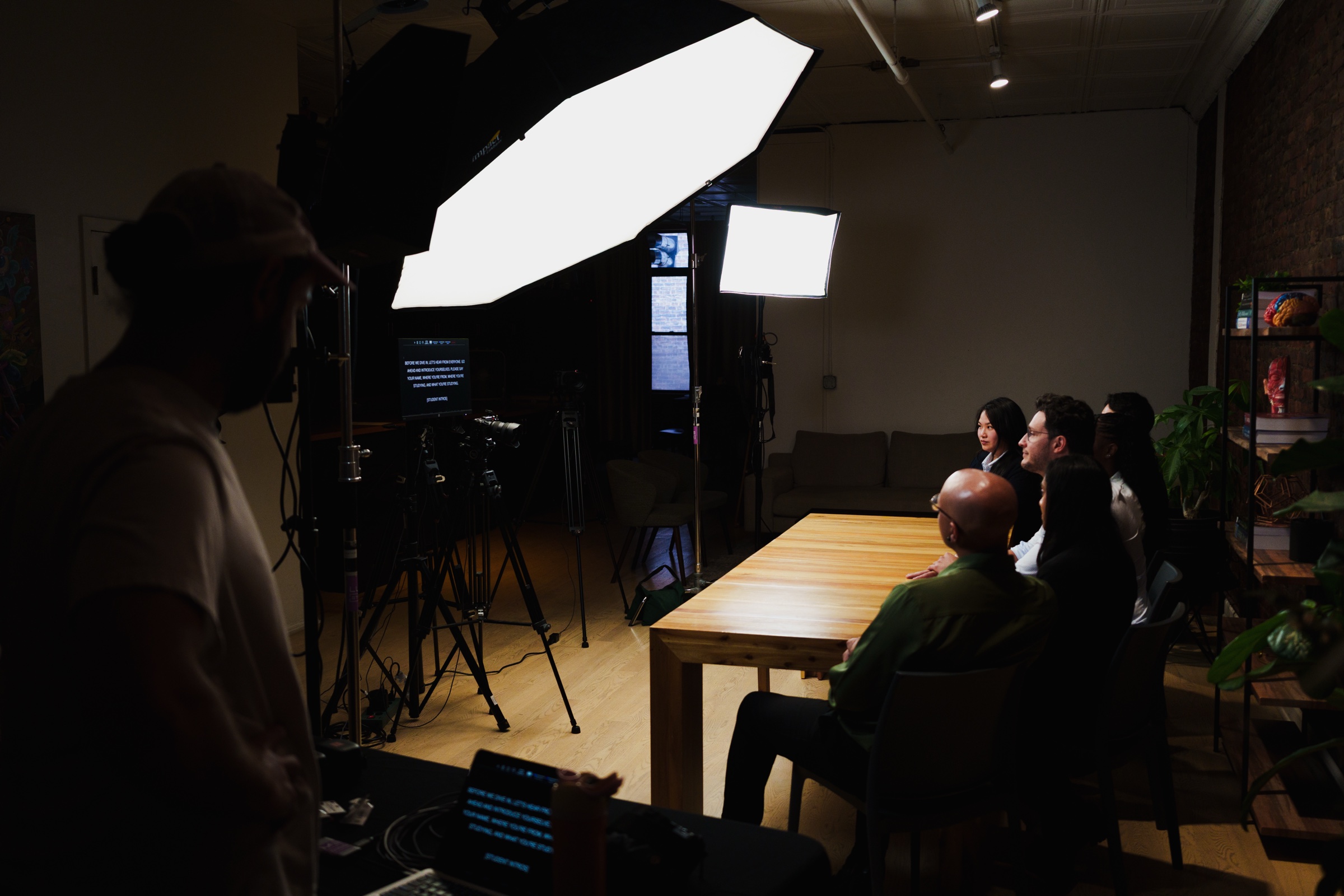 Silhouetted crew watch from behind lights and monitors while the guests sit lit at the table