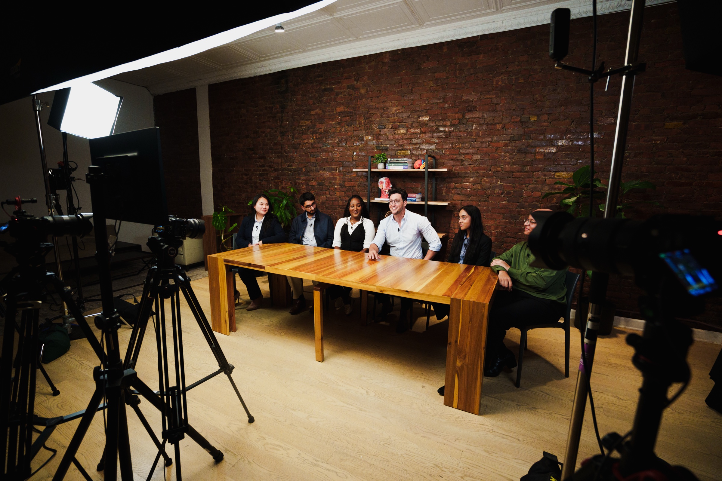 Six people sit along the table facing a row of tripods and softboxes set across the floor