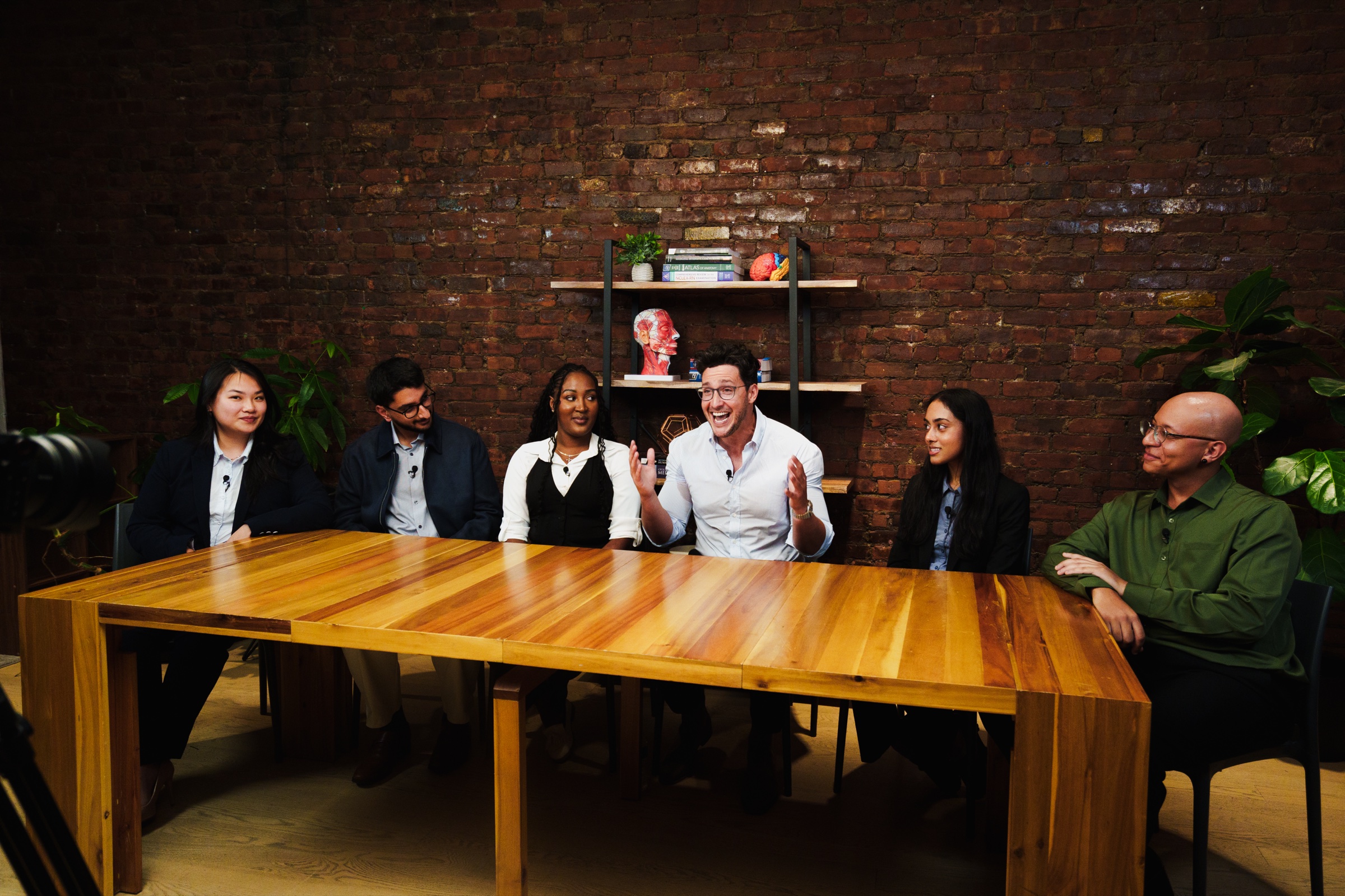 Six people at the table laugh as the man in the center speaks with his hands raised