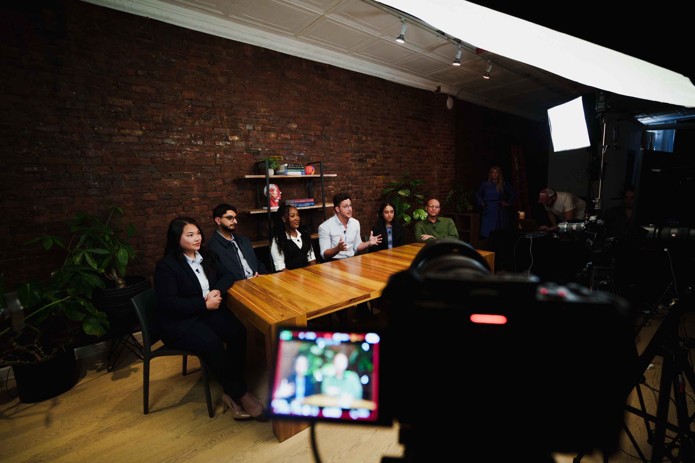 The group sits at the table seen past a camera whose screen shows the same shot