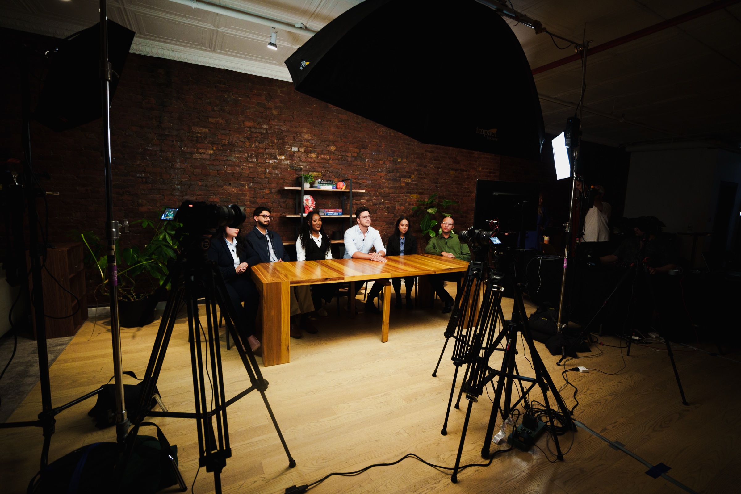 Two cameras on tripods face the table where six people sit under a large black umbrella light