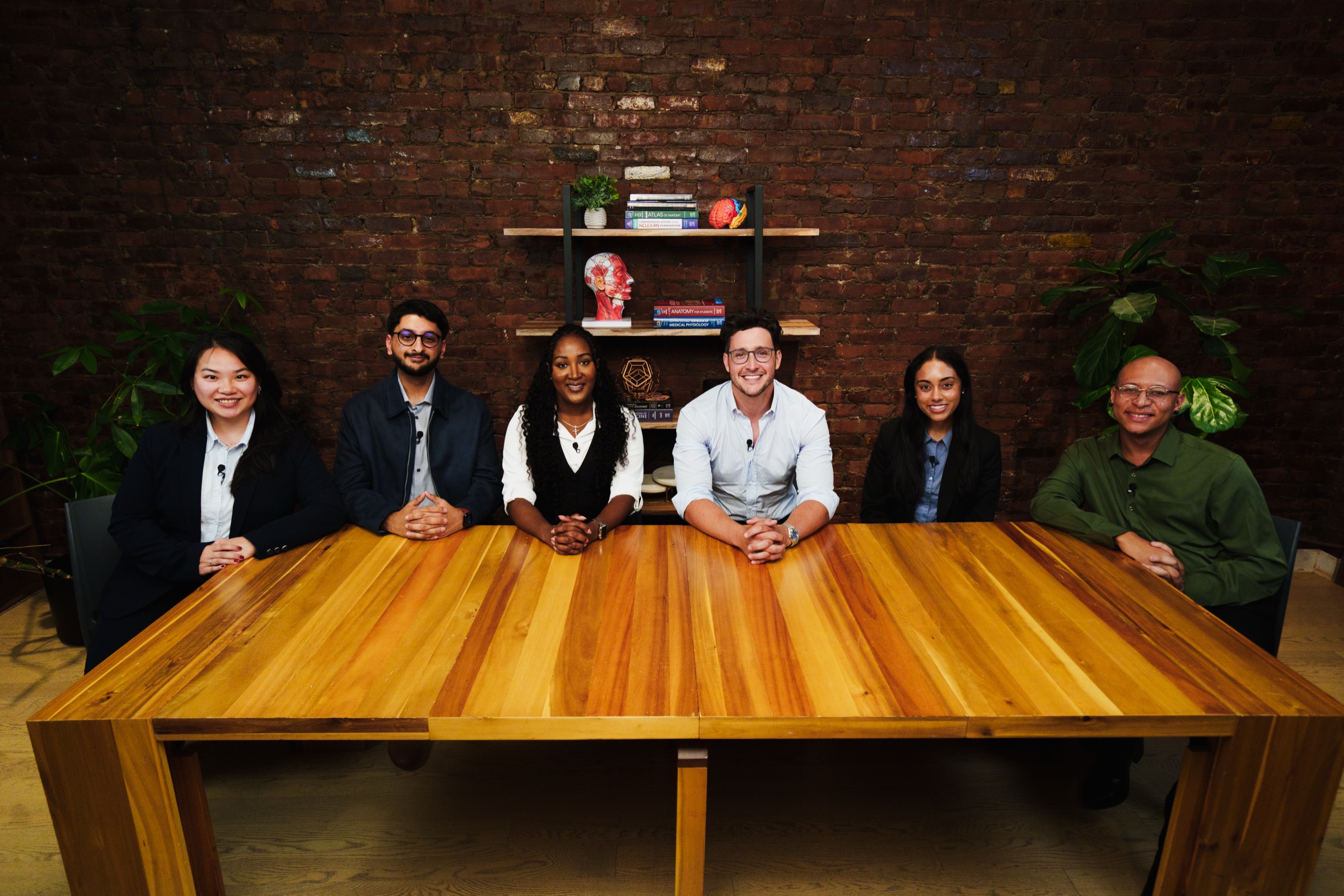 Six people sit shoulder to shoulder behind the wooden table, smiling at the camera against brick