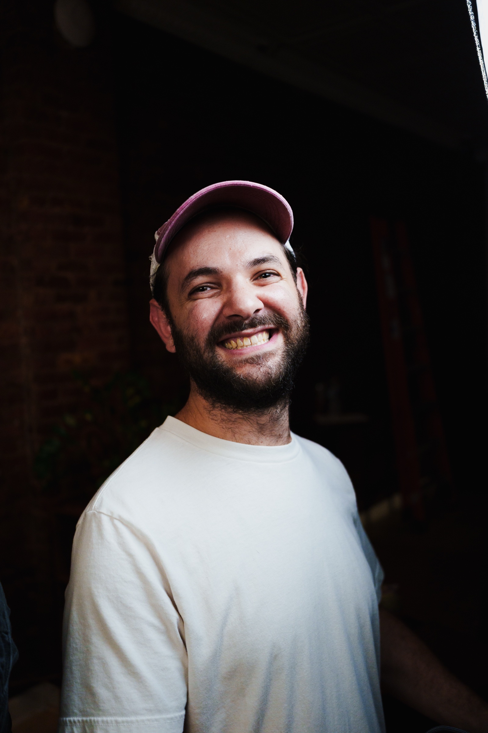 Doctor Mike smiles in a pink cap and cream tee shirt, brick wall dark behind him