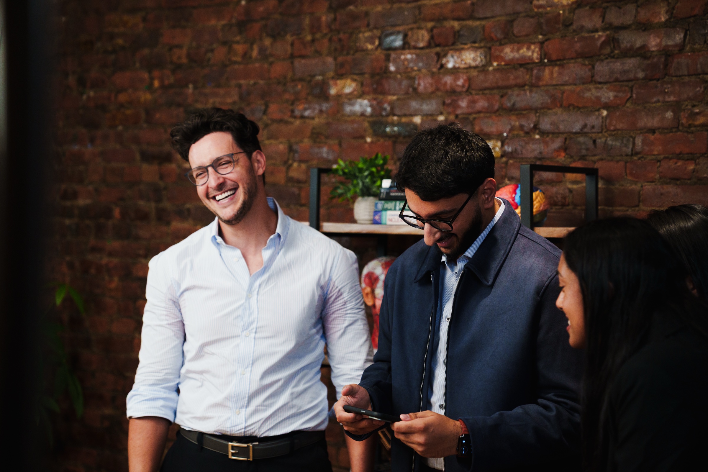 Two men laugh as one checks a phone, a woman beside them by the brick wall